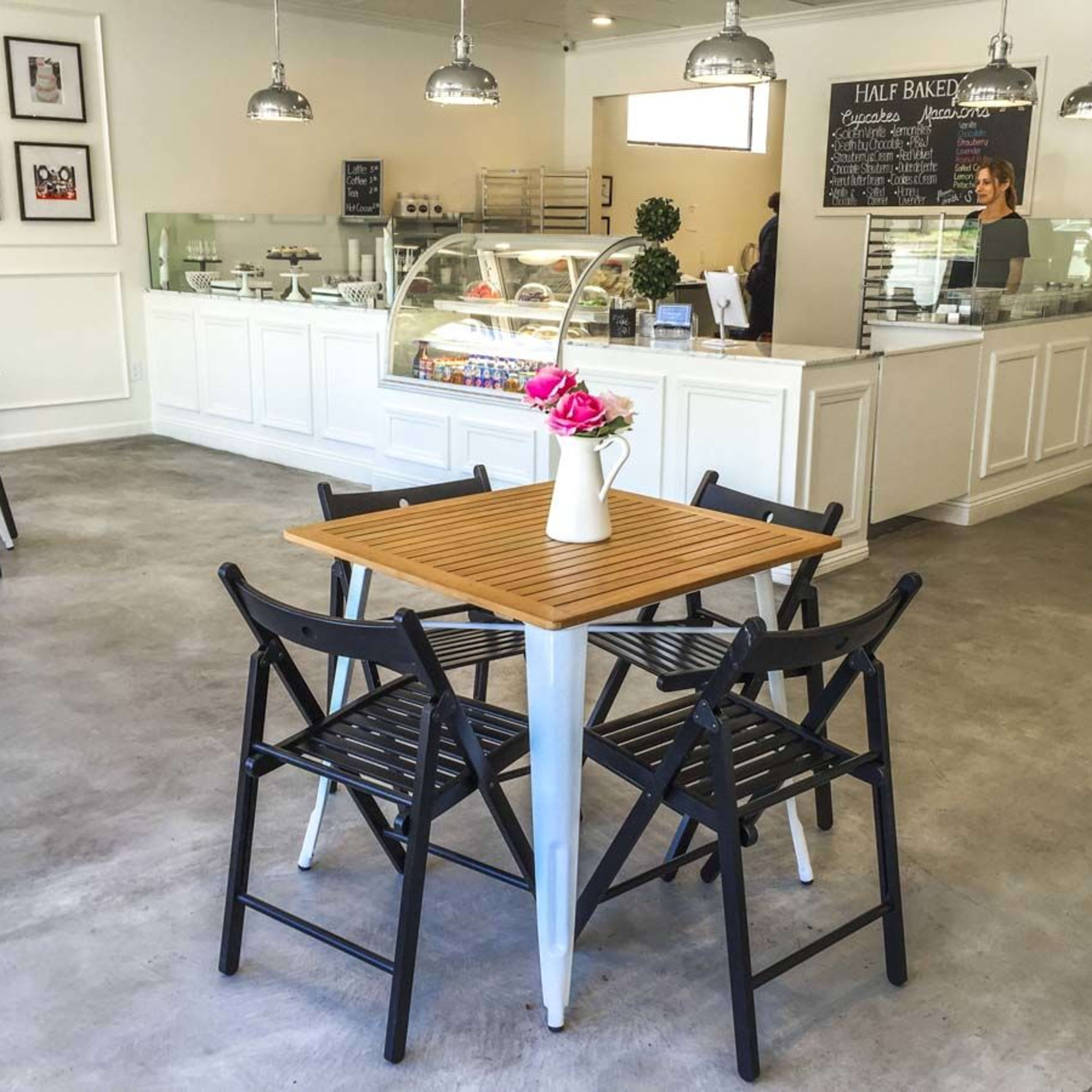 A bright and modern cafe with a wooden table and black folding chairs arranged in the center, complemented by a glass display case of desserts in the background.