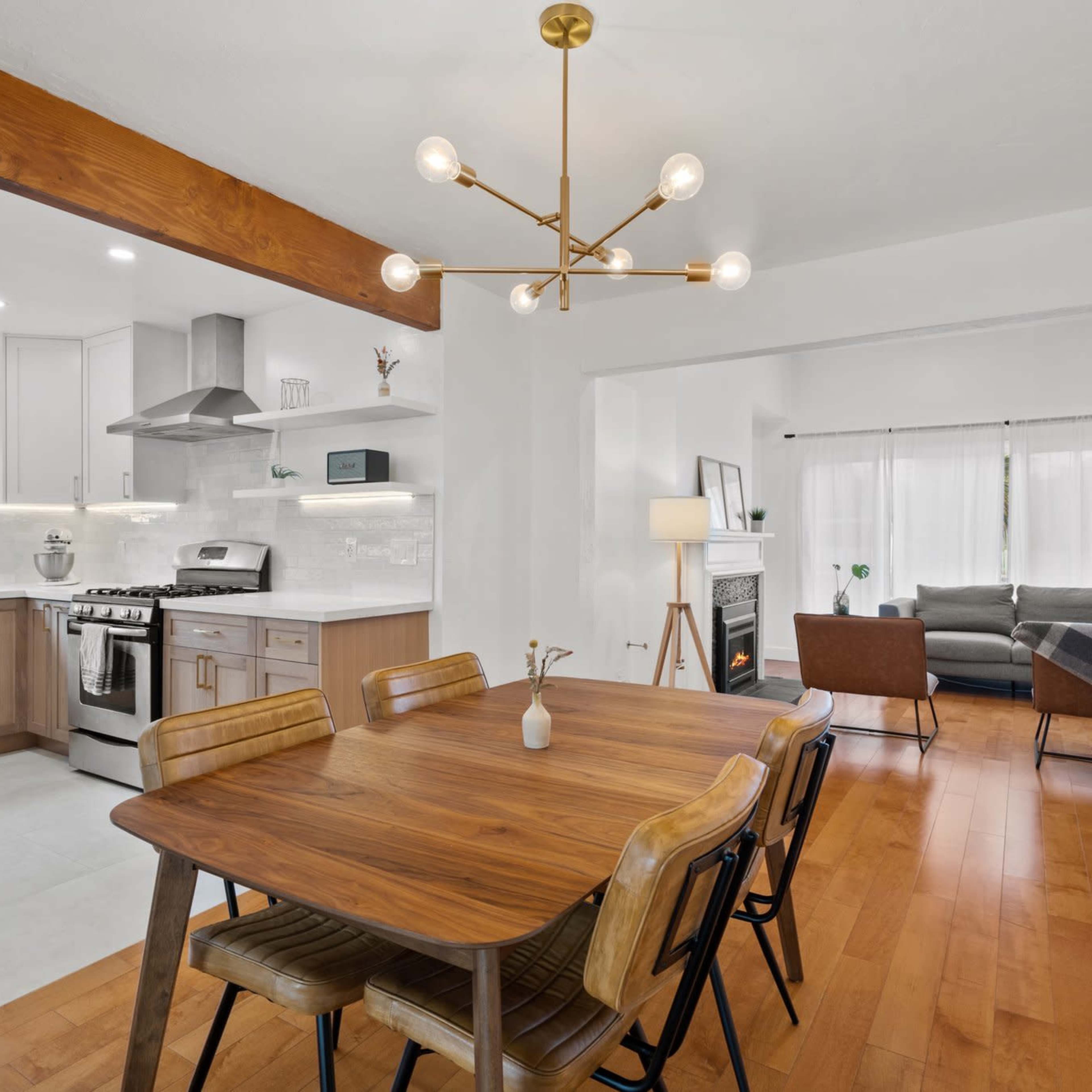 A modern kitchen and dining area featuring a wooden dining table, sleek appliances, and a living room visible in the background with a couch and large windows.