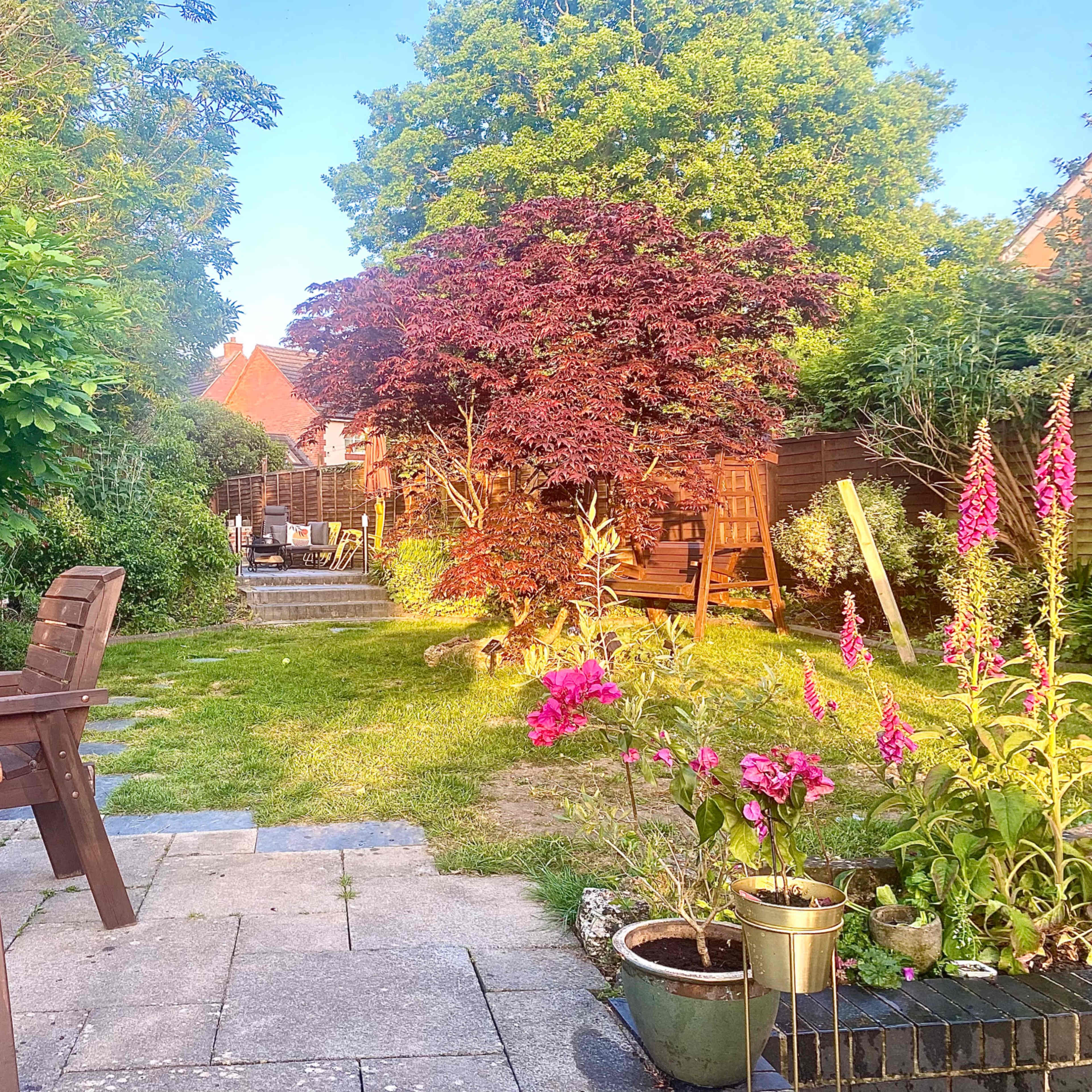 A garden scene features a pathway lined with flowers, wooden seating areas, and lush greenery, leading to a distant seating arrangement.
