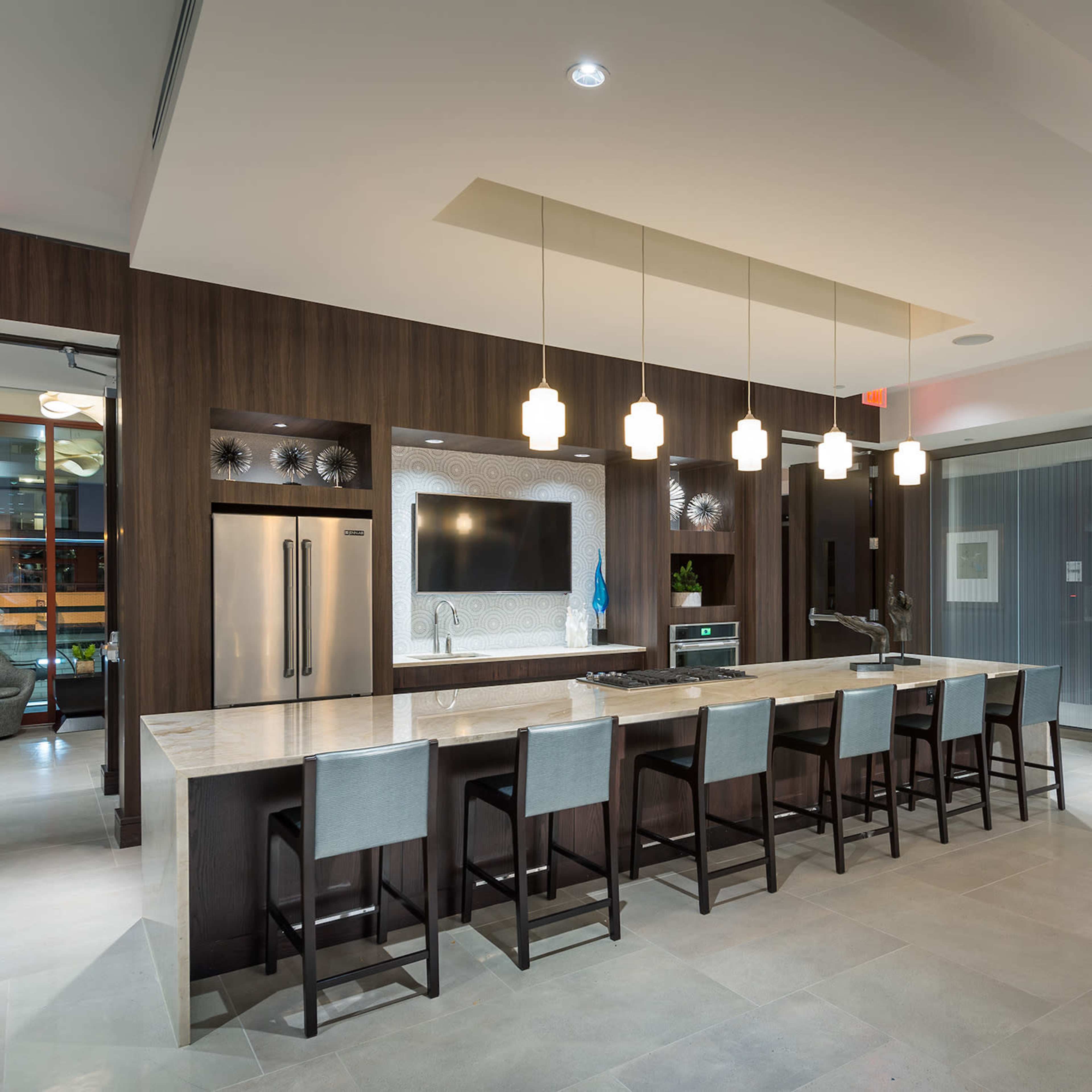 A modern lobby features a gray sofa, marble surfaces, a large television, and a bar area with stools.