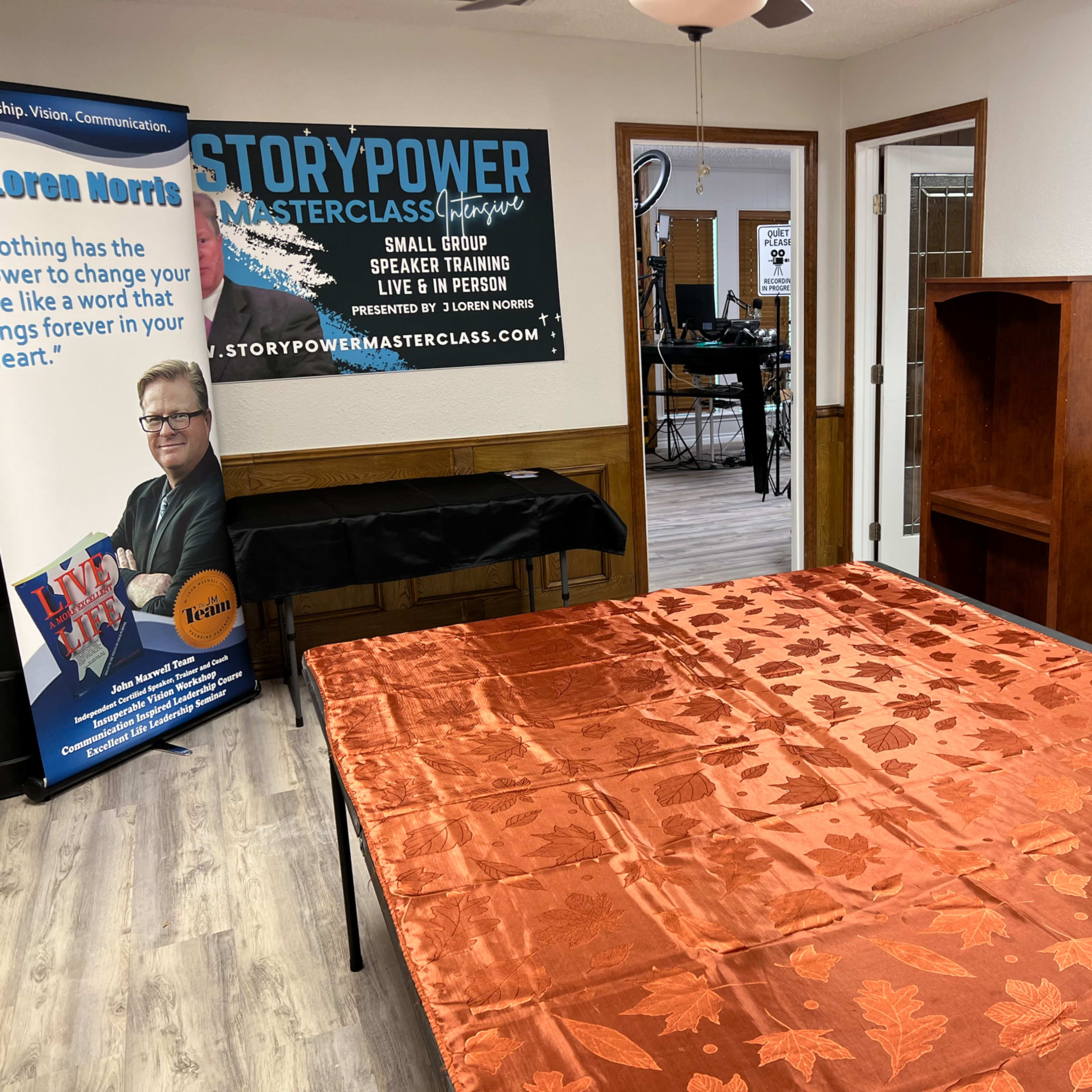 A training room with a large orange table covered in a leaf-patterned tablecloth, a banner featuring a speaker's photo, and a wooden shelf against the wall.