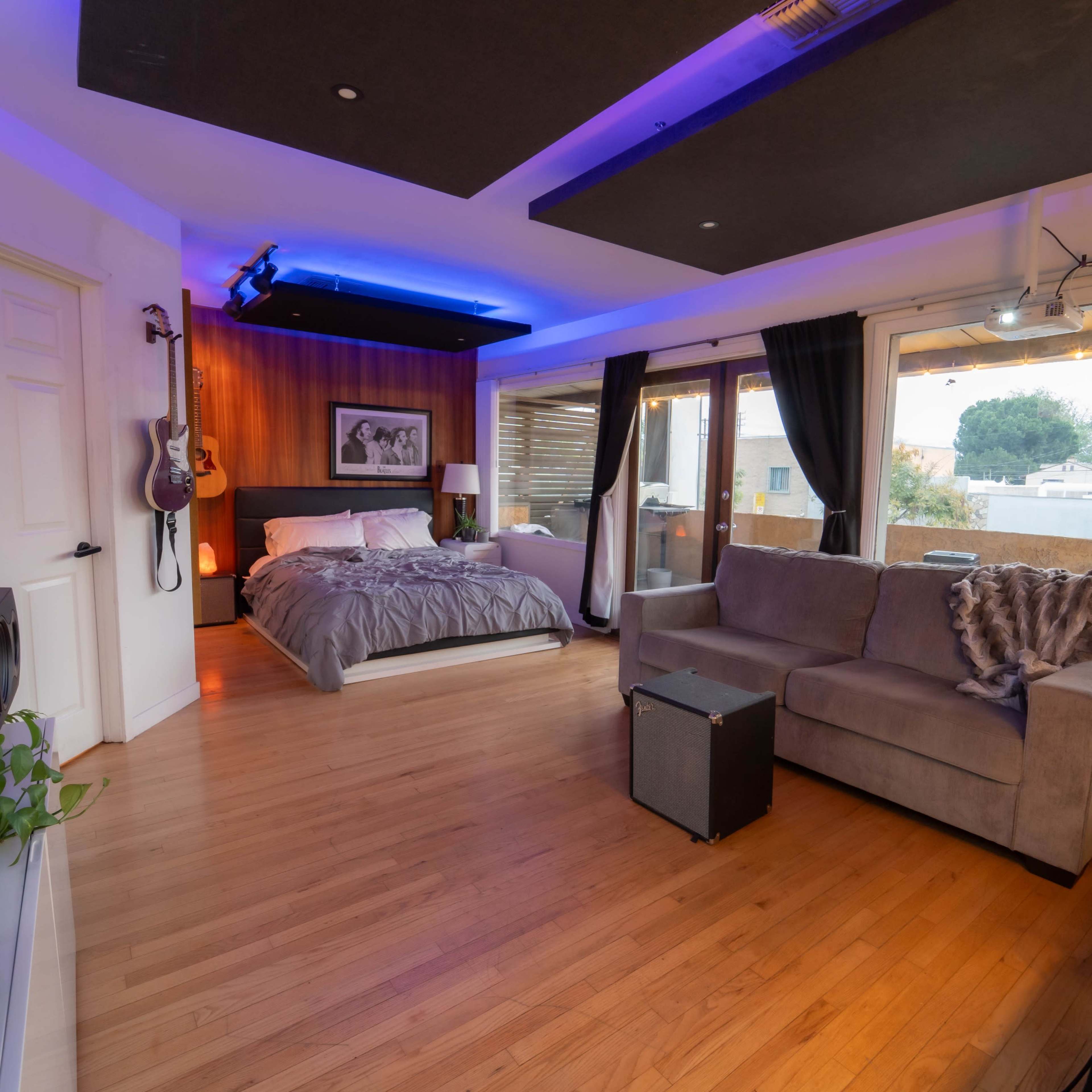 A modern studio apartment featuring a bed, a gray sofa, large windows, and a small audio setup.