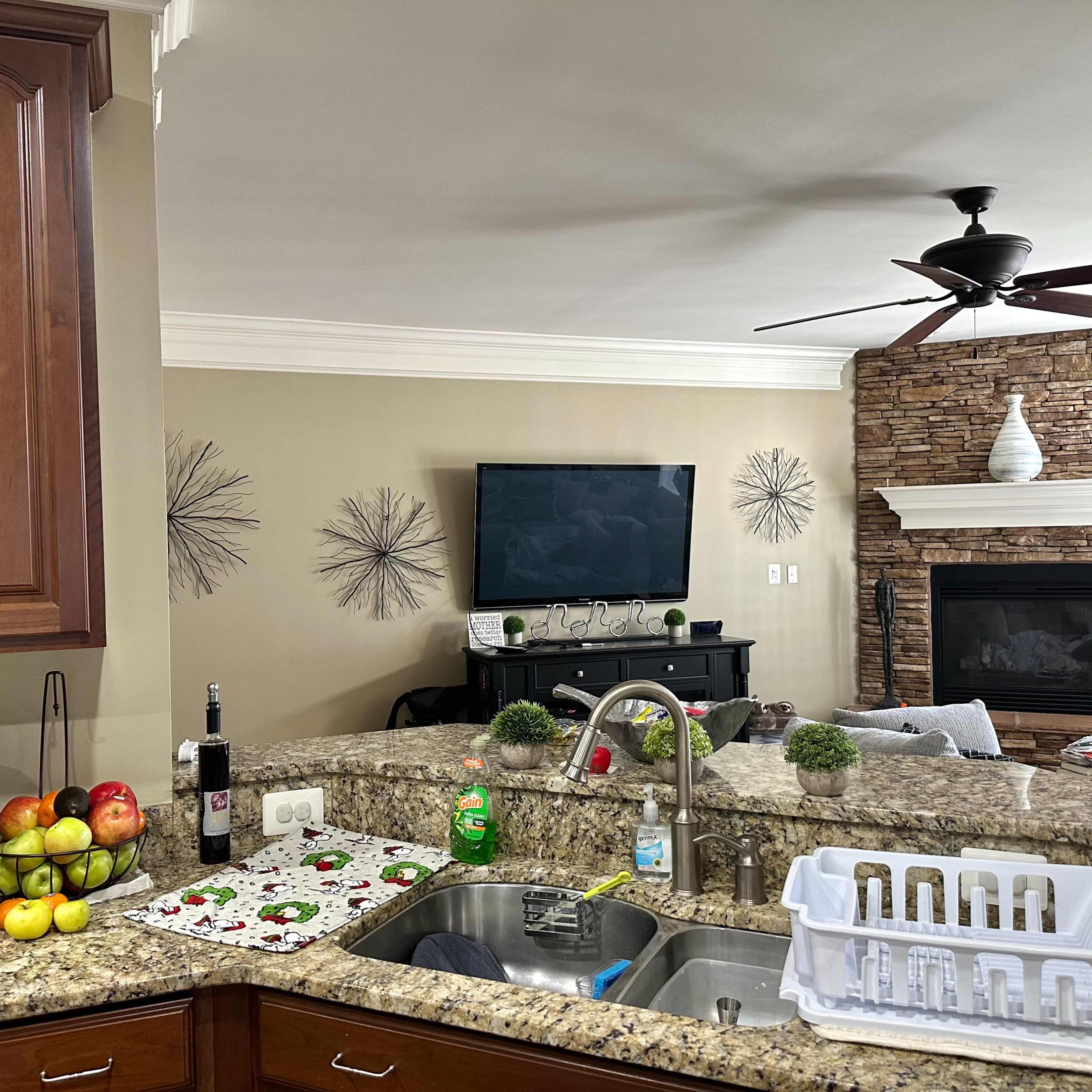A kitchen with wooden cabinets and granite countertops, featuring a sink and a view into a living area that includes a TV and a stone fireplace.