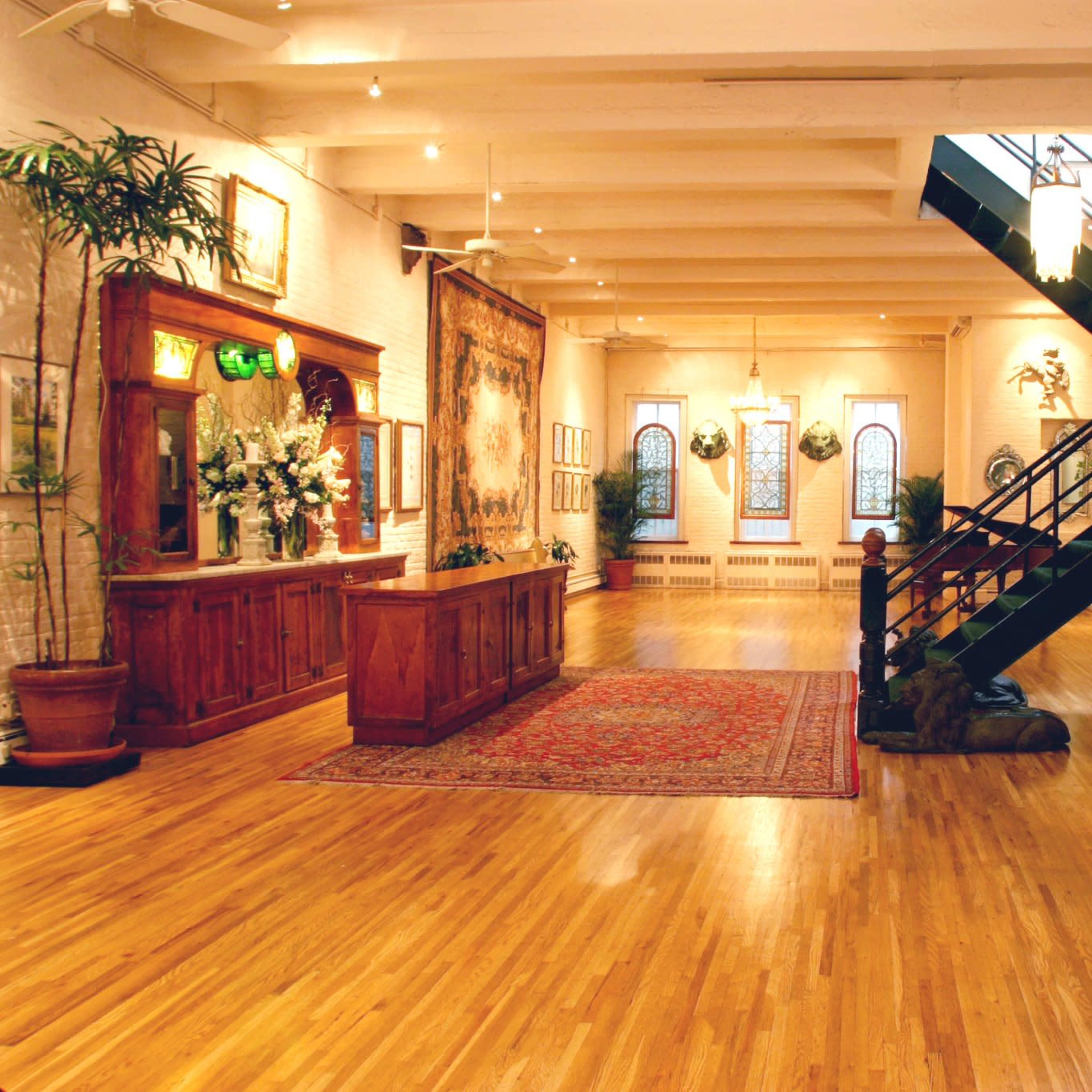 A spacious gallery with hardwood floors, a staircase leading to an upper level, and a display of artworks and plants along the walls.
