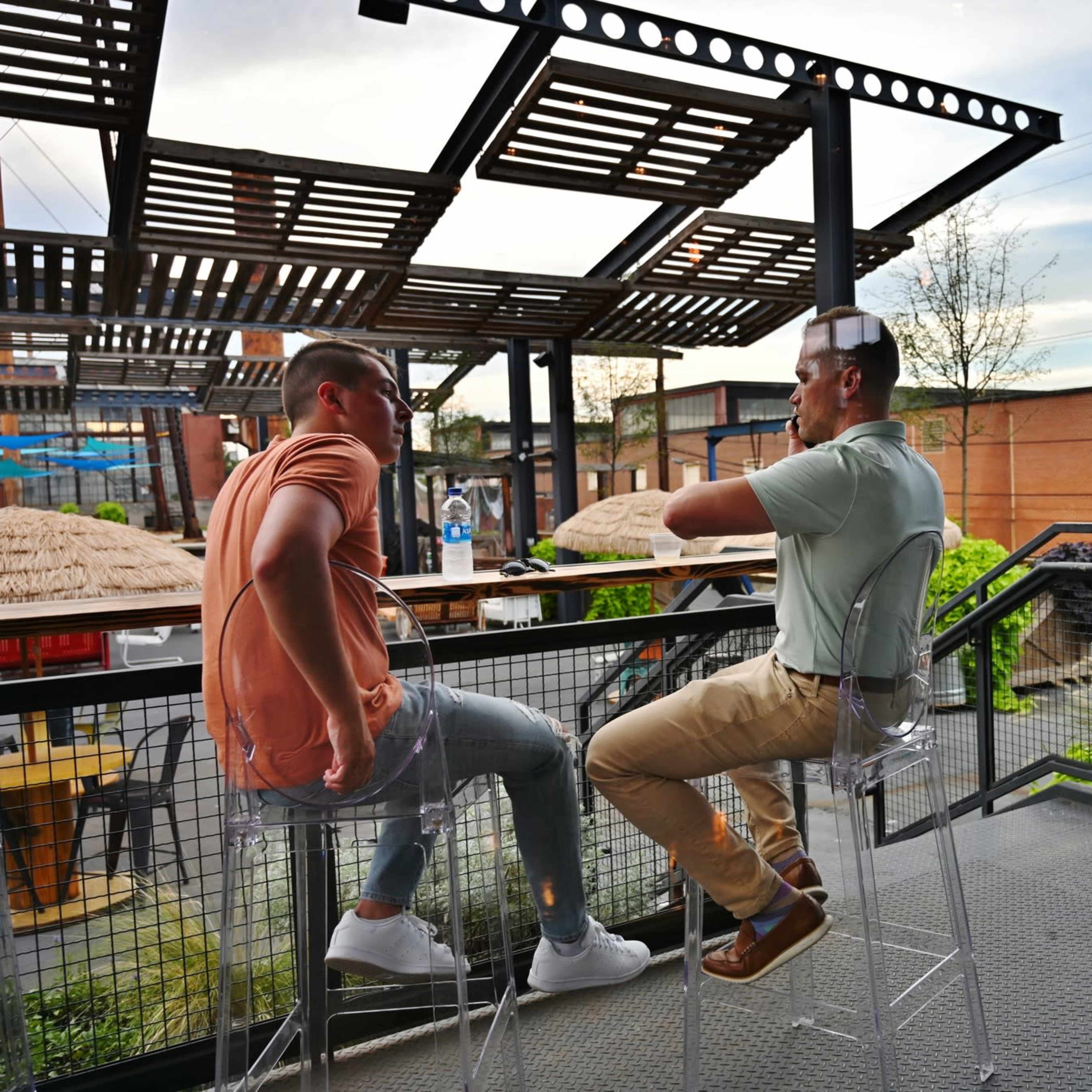 Two men sit on transparent stools at a bar overlooking an outdoor area, while another couple engages in conversation at a nearby table.