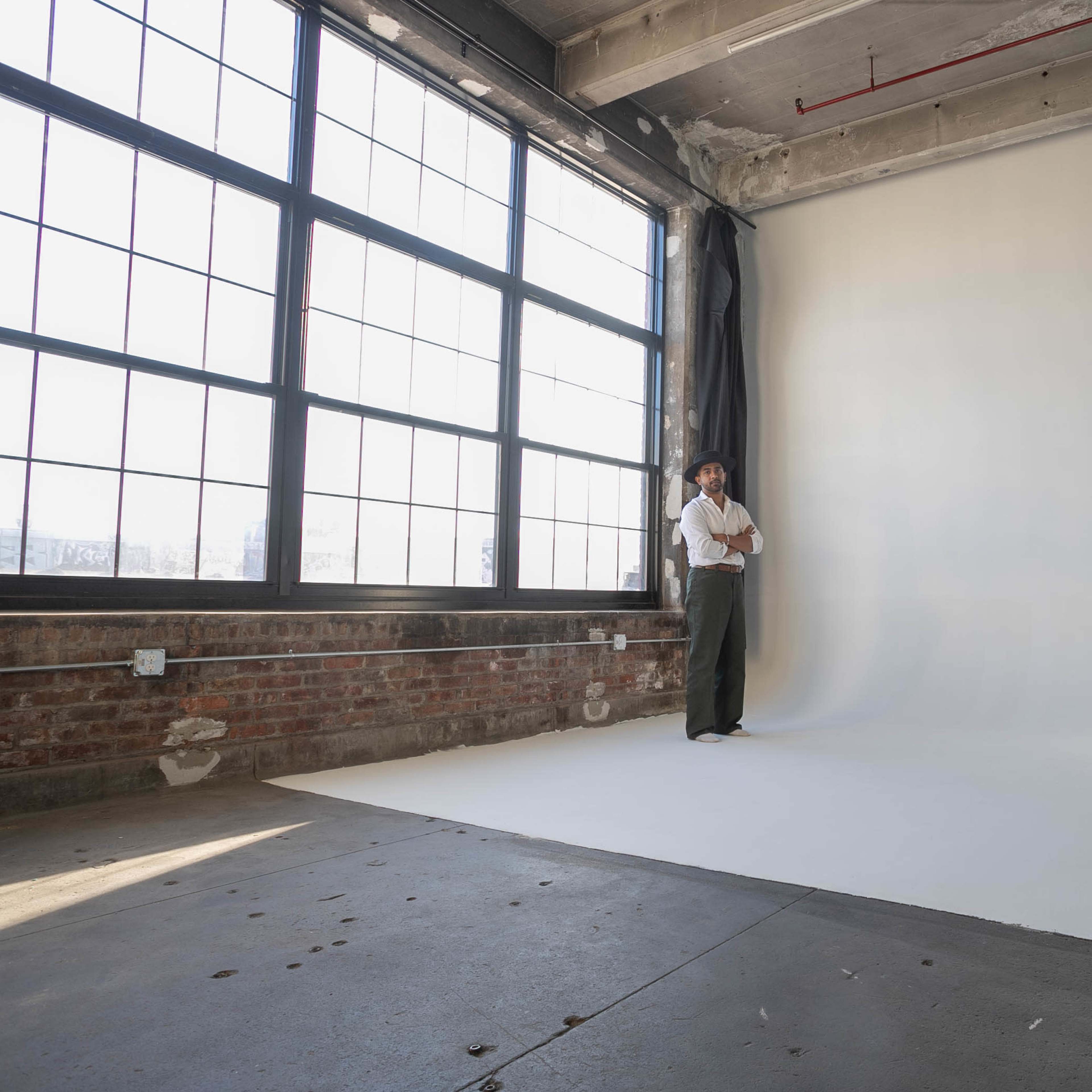 A person stands in a spacious, well-lit photo studio with large windows and a plain white backdrop.
