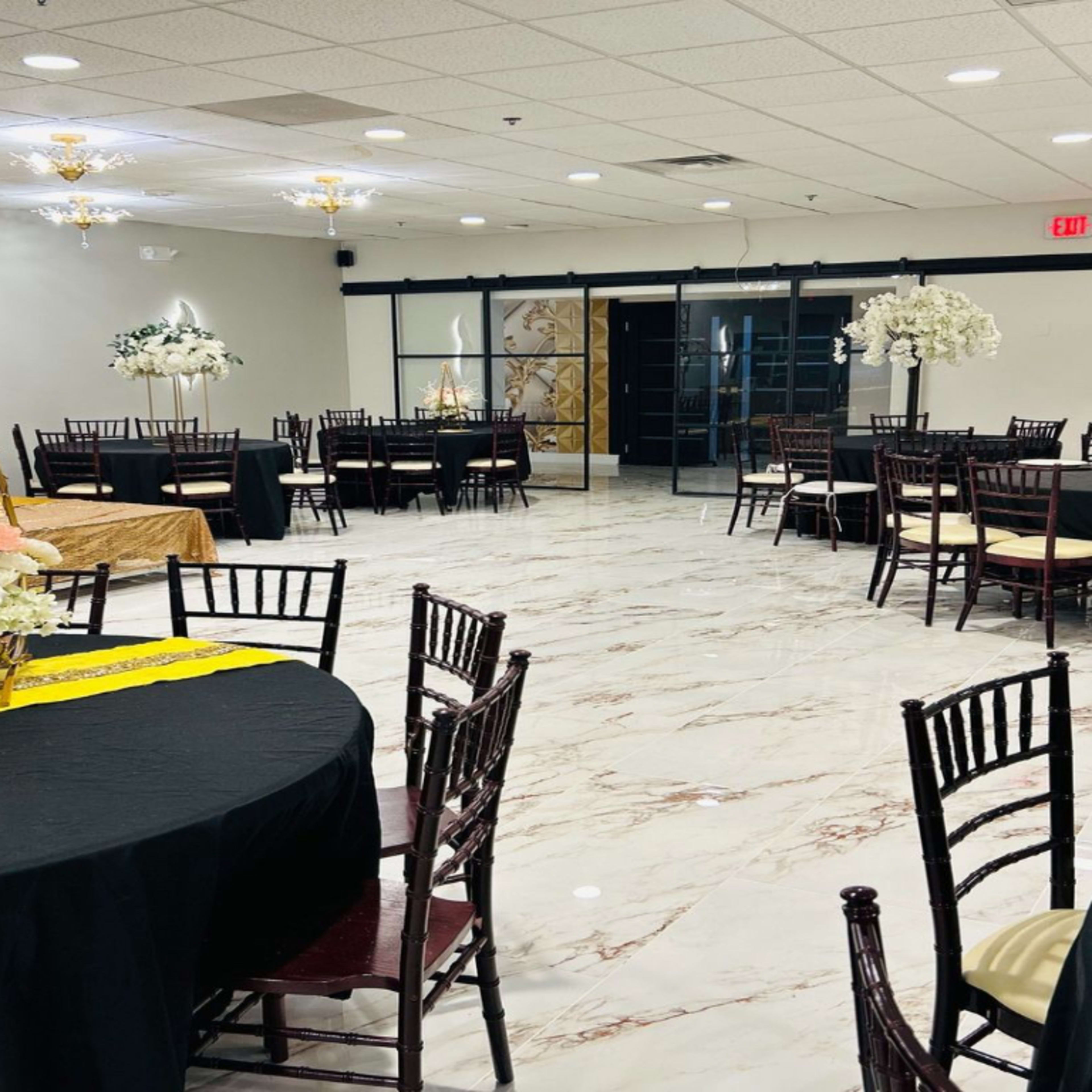 A spacious event venue with tables set for a gathering, featuring black and gold decor and flower arrangements.