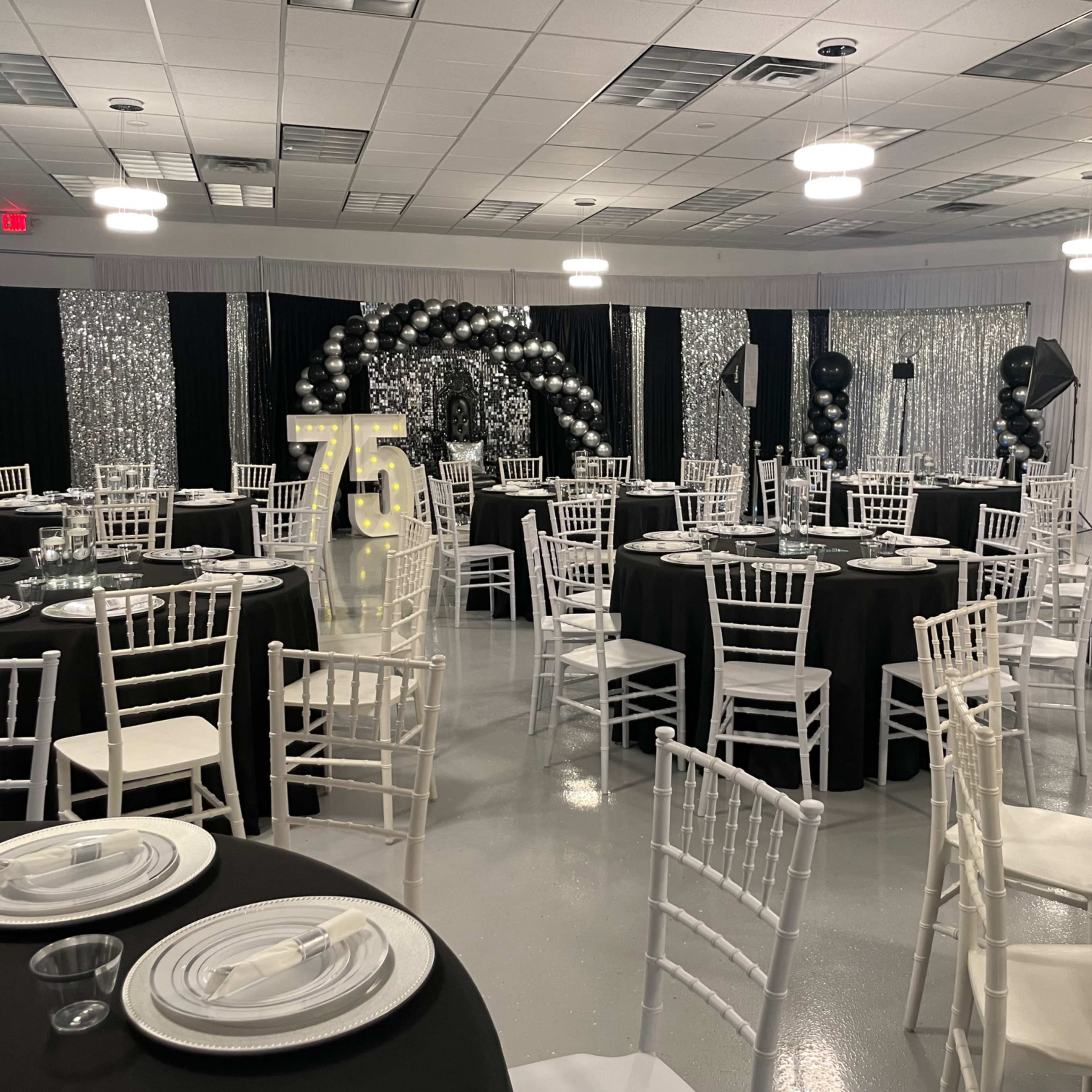 A decorated banquet hall set for a event, featuring black and white tables, a large "75" sign, and balloon arch decorations.
