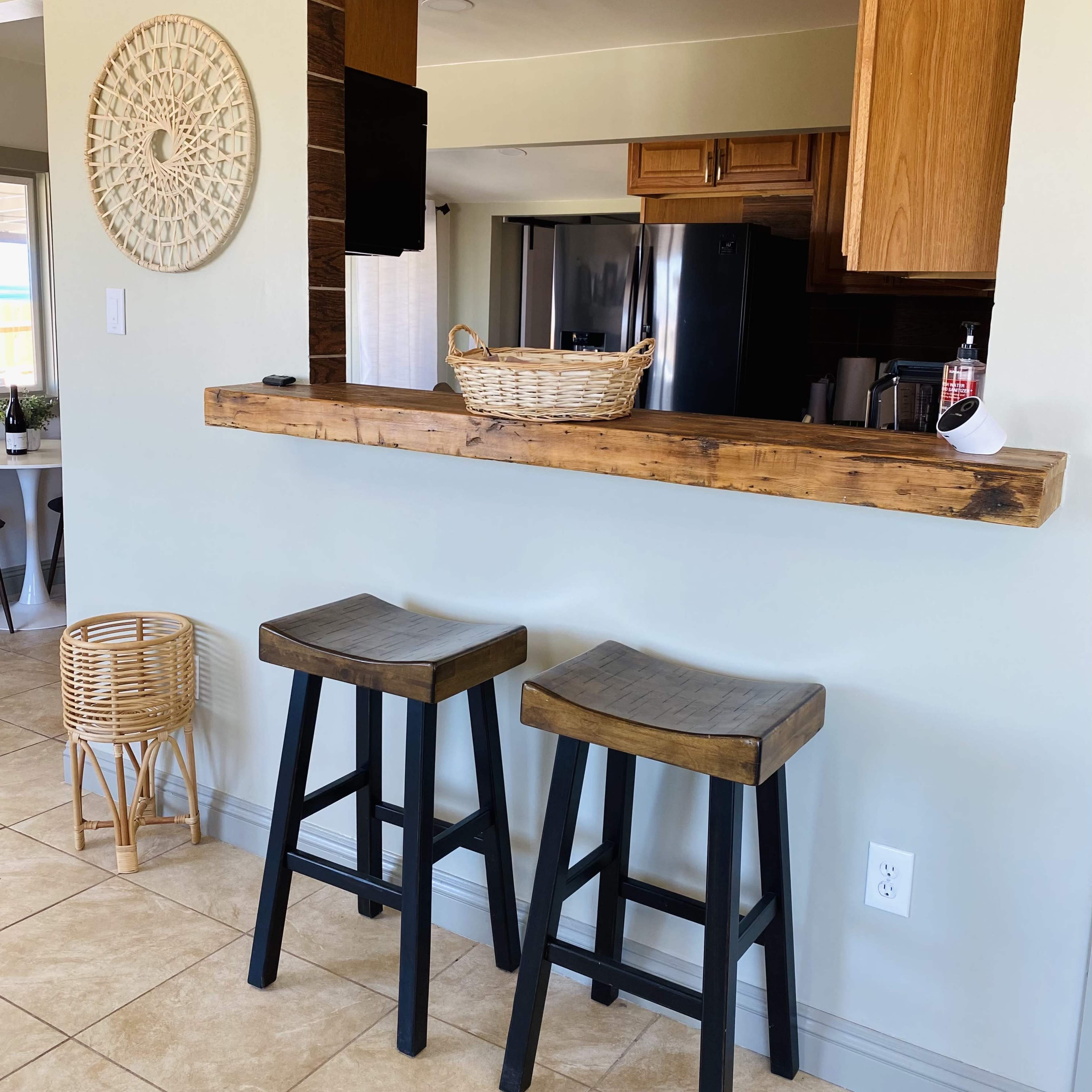 Two wooden bar stools are positioned next to a flat wooden countertop, with a woven basket on top, against a backdrop of a kitchen area.