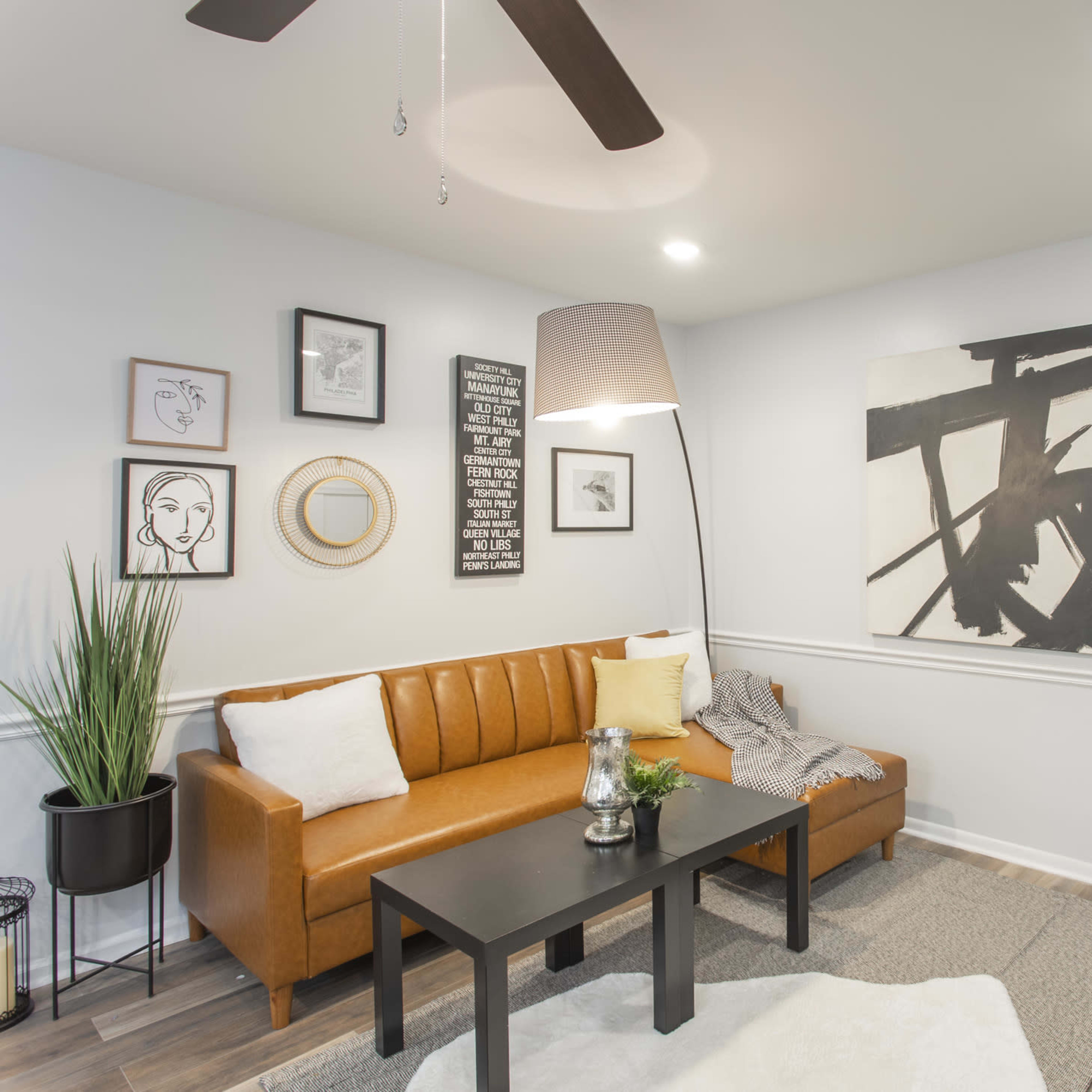 A modern living room features a brown leather sofa, a black coffee table, artwork on the walls, and a gray chair, all arranged with a light rug on a wooden floor.