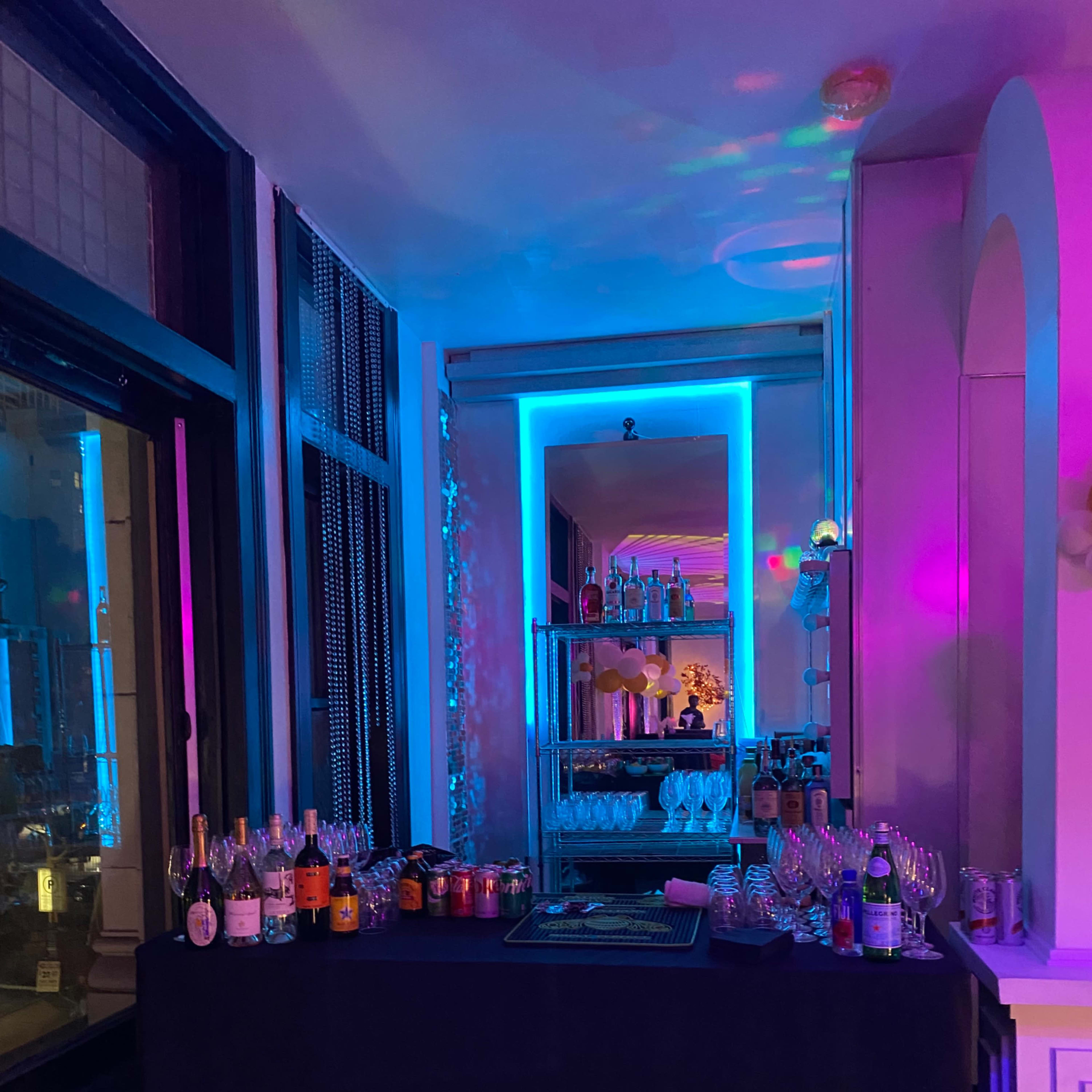 A bar area is set up with a variety of bottles and glasses, illuminated by colorful lighting against a backdrop of shelves filled with more liquor and glassware.