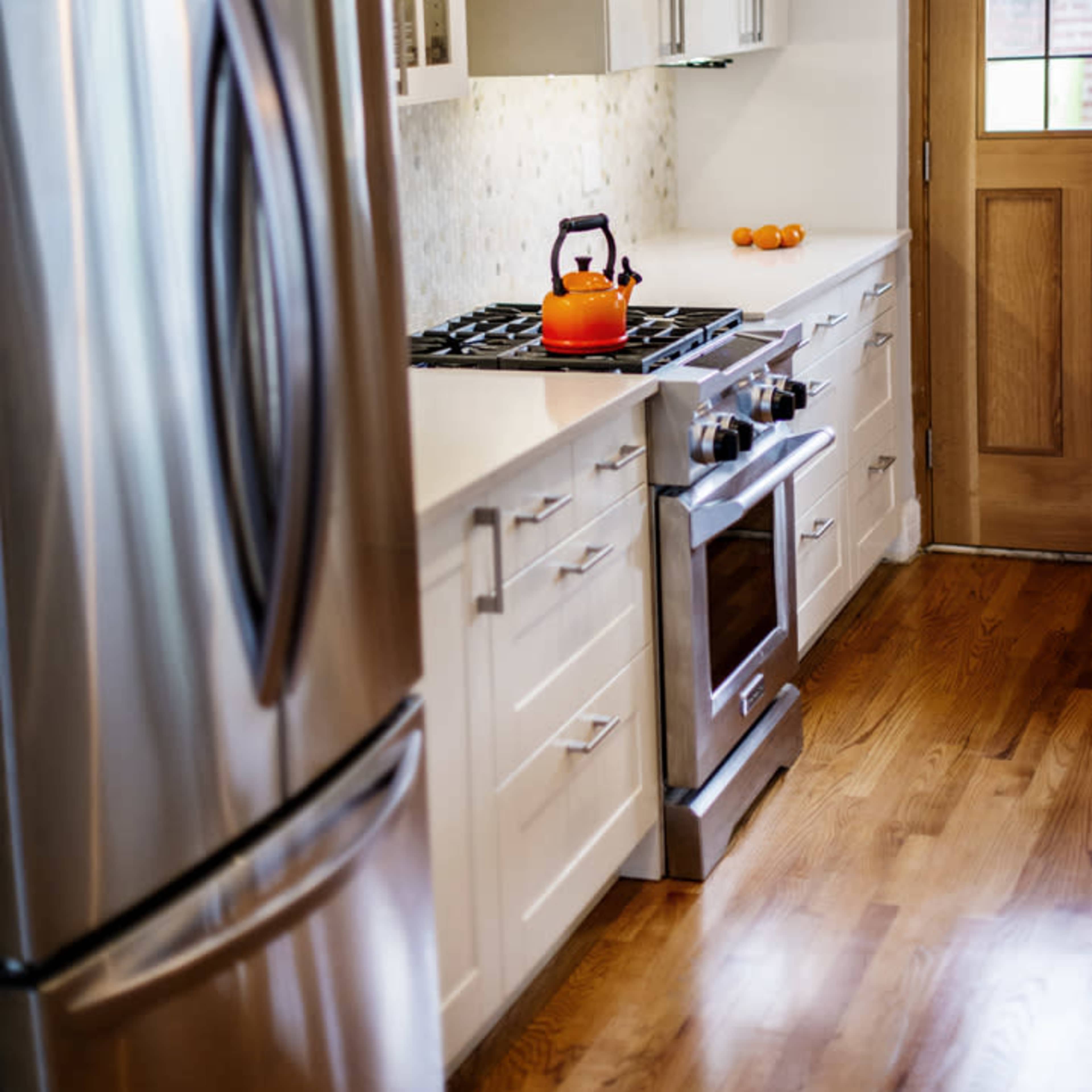 A modern kitchen with stainless steel appliances, white cabinetry, and a wooden door, featuring an orange kettle on the stove and three oranges on the counter.