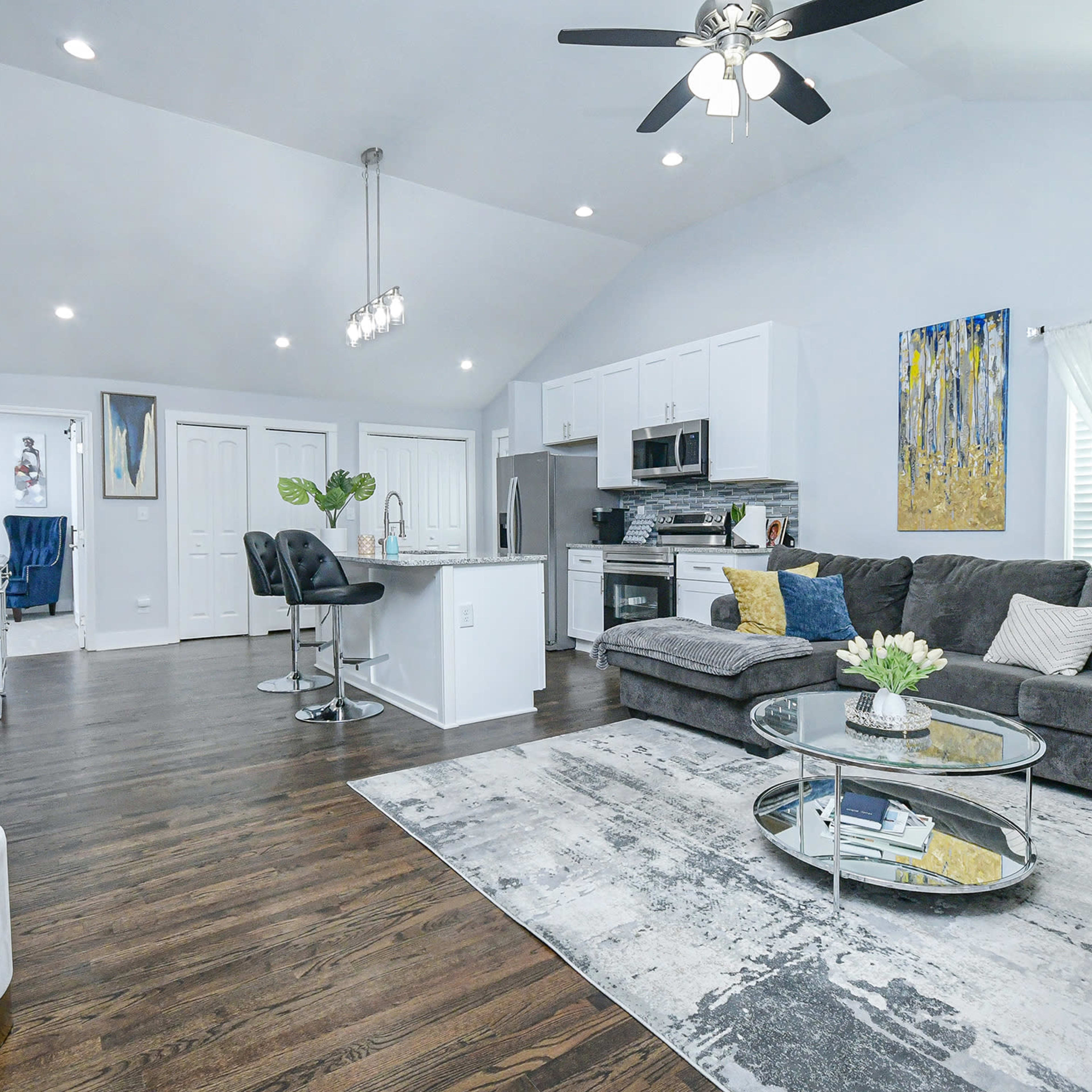A modern living room with a sectional sofa, a round coffee table, a kitchen area, and large windows allowing natural light to enter.