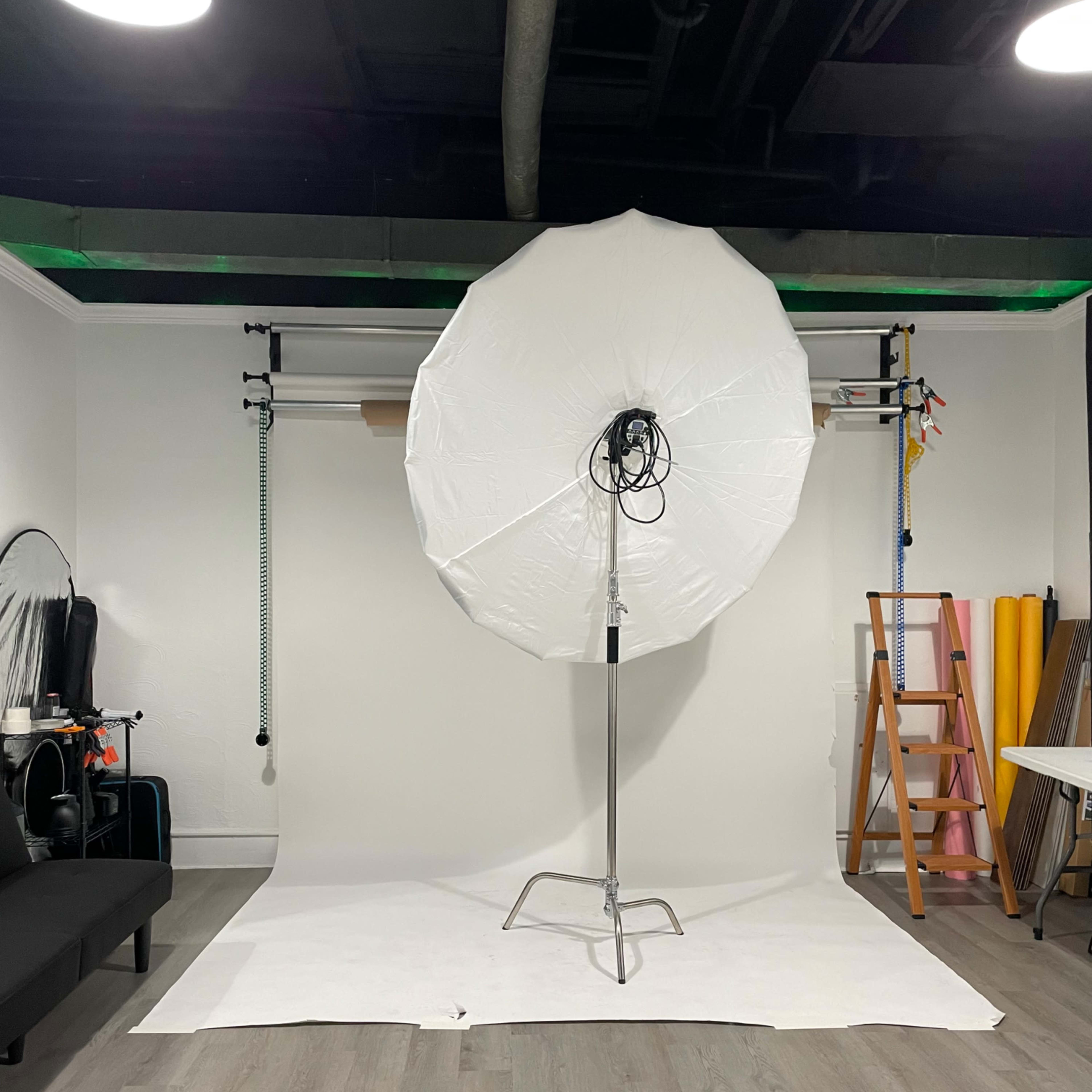 A photography studio with an umbrella softbox positioned in front of a plain backdrop, surrounded by various studio equipment and a ladder.