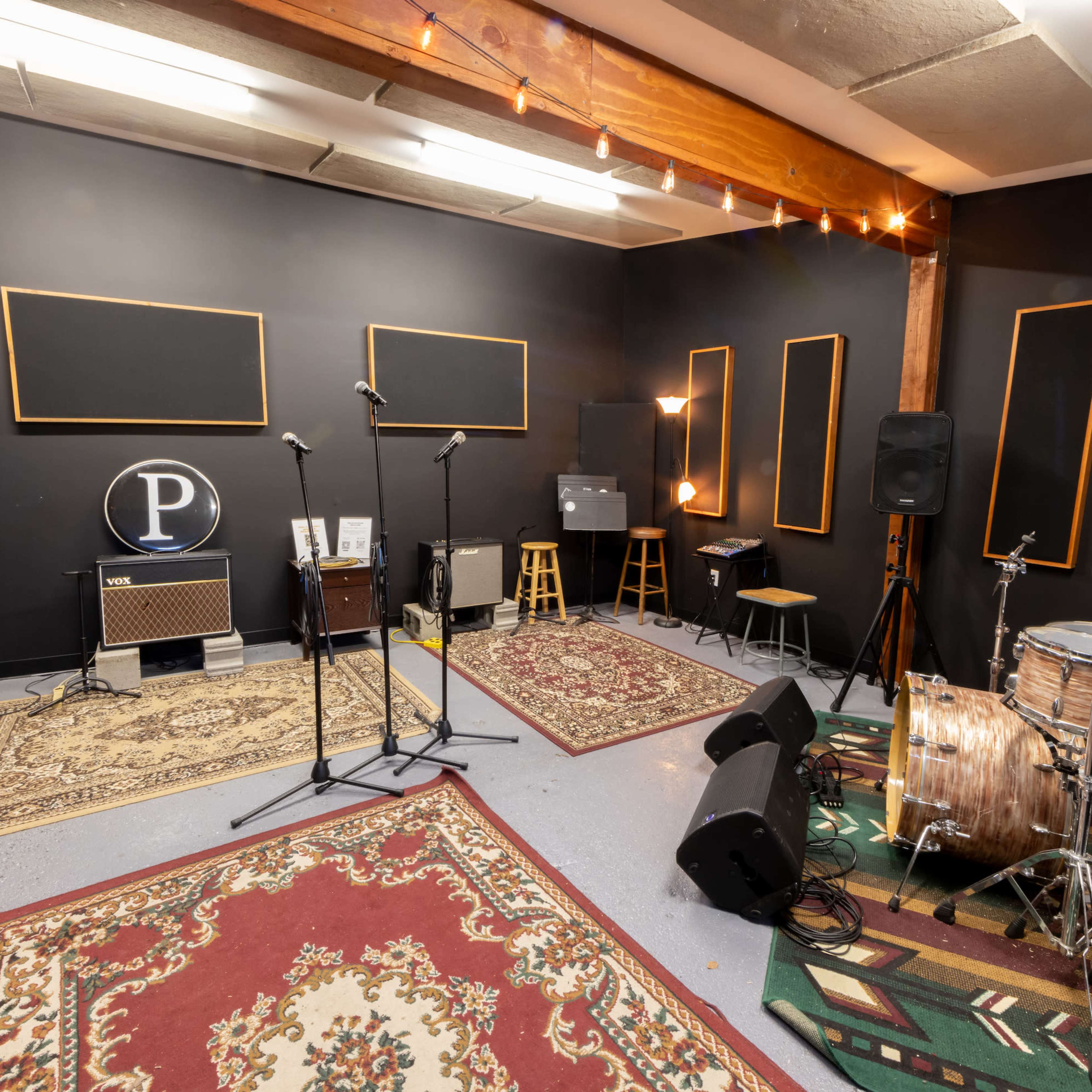 A music rehearsal space featuring two microphone stands, a drum set, amplifiers, and various seating arrangements against a backdrop of black walls and area rugs.