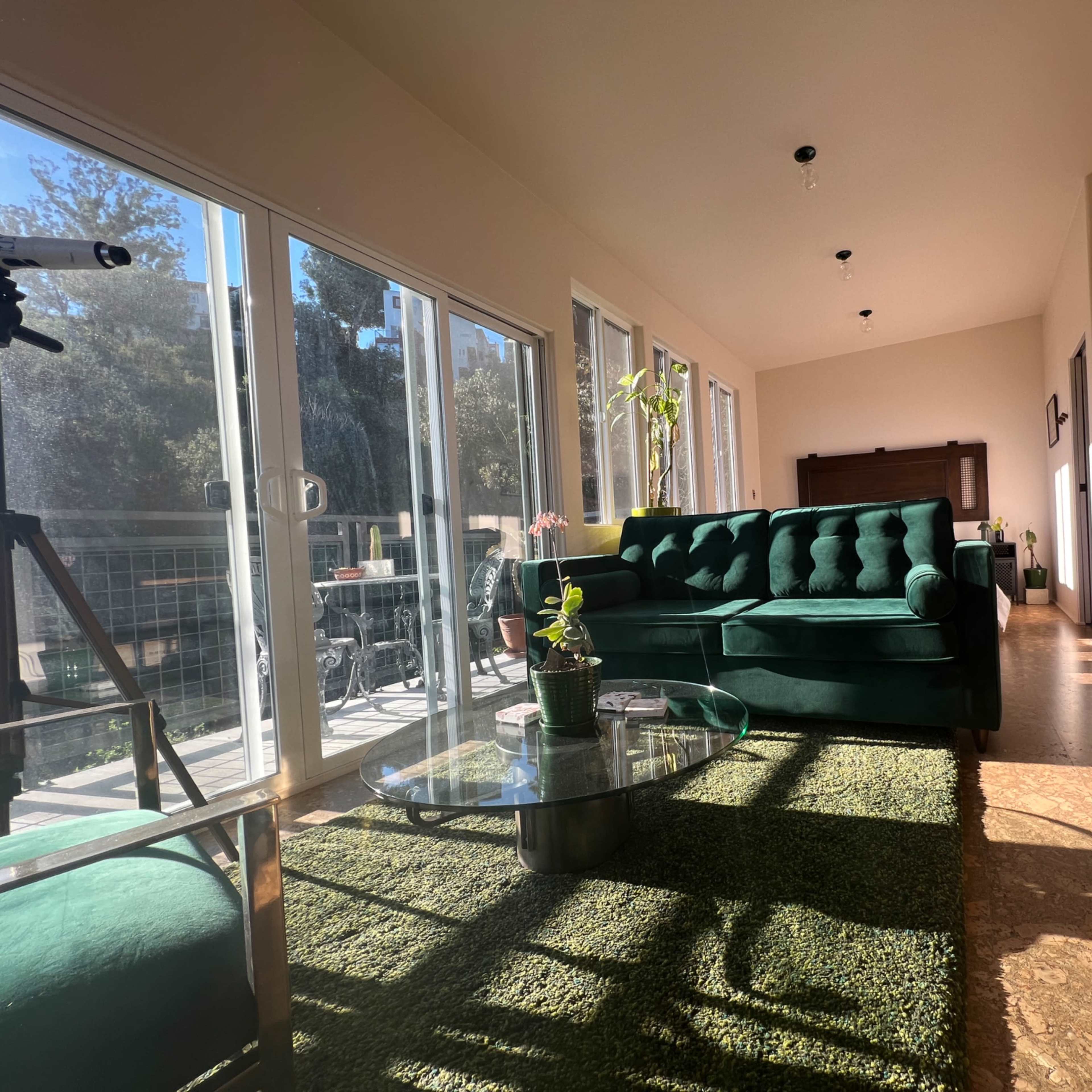 A well-lit living room featuring a green tufted sofa, a glass coffee table, and large windows displaying an outdoor view.