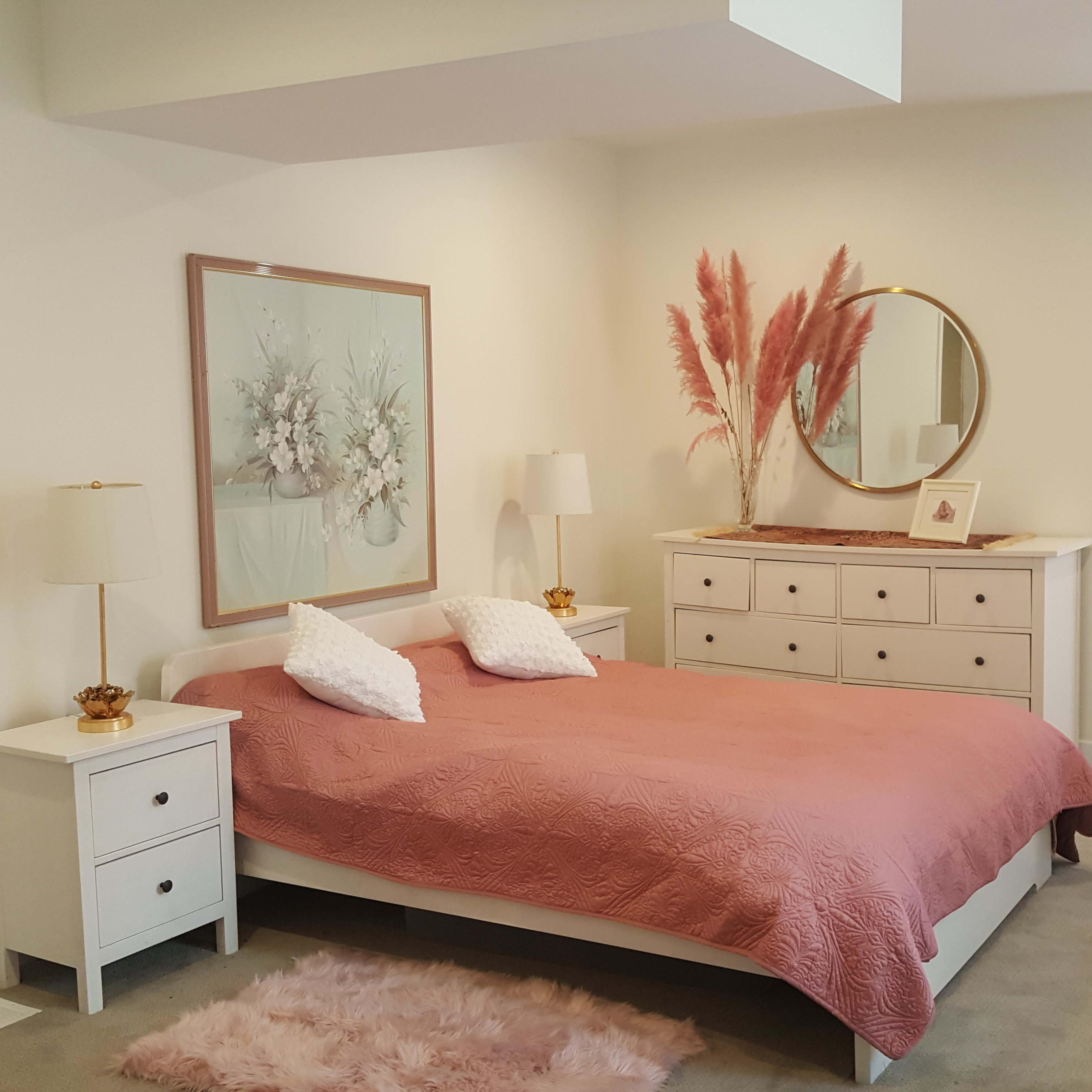 A neatly arranged bedroom features a bed with a pink quilt, bedside tables with lamps, a dresser, and decorative items, including a round mirror and pampas grass.