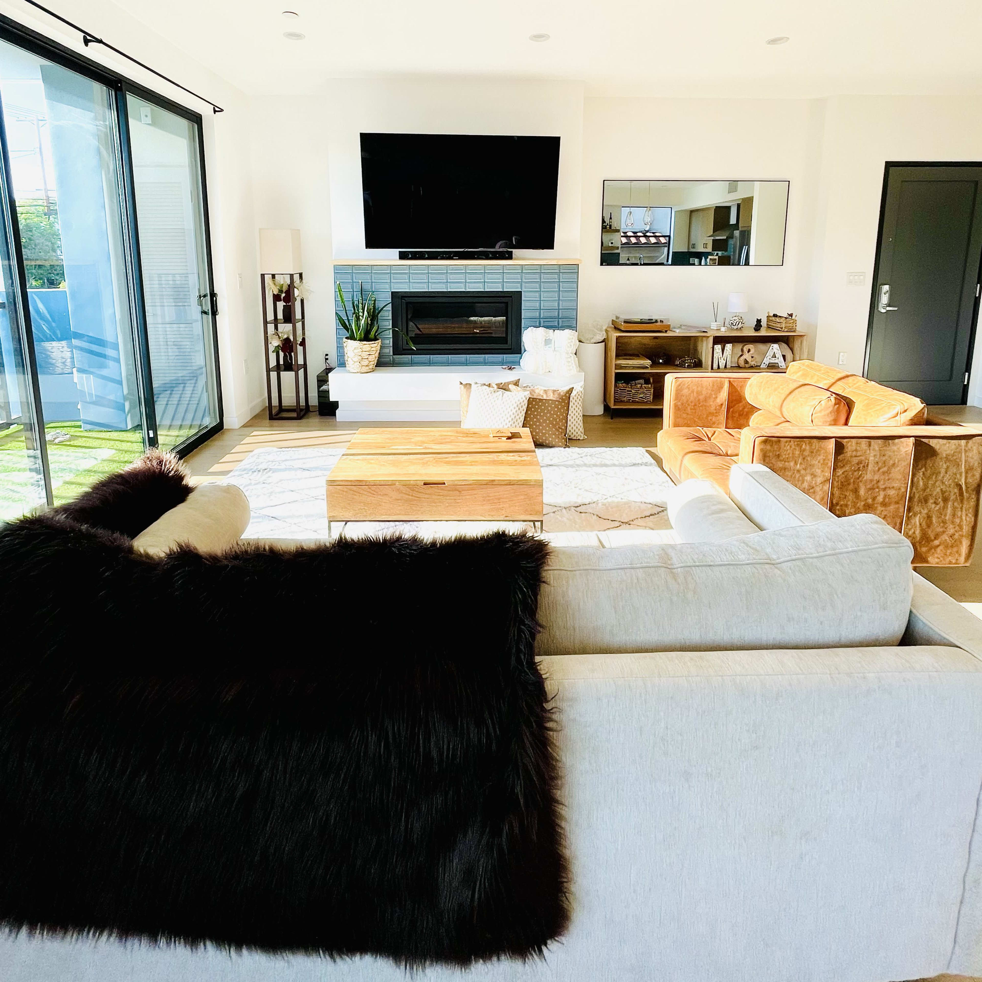 A modern living room features a television mounted above a fireplace, a wooden coffee table, and a mix of seating including a white sofa and an orange chair.