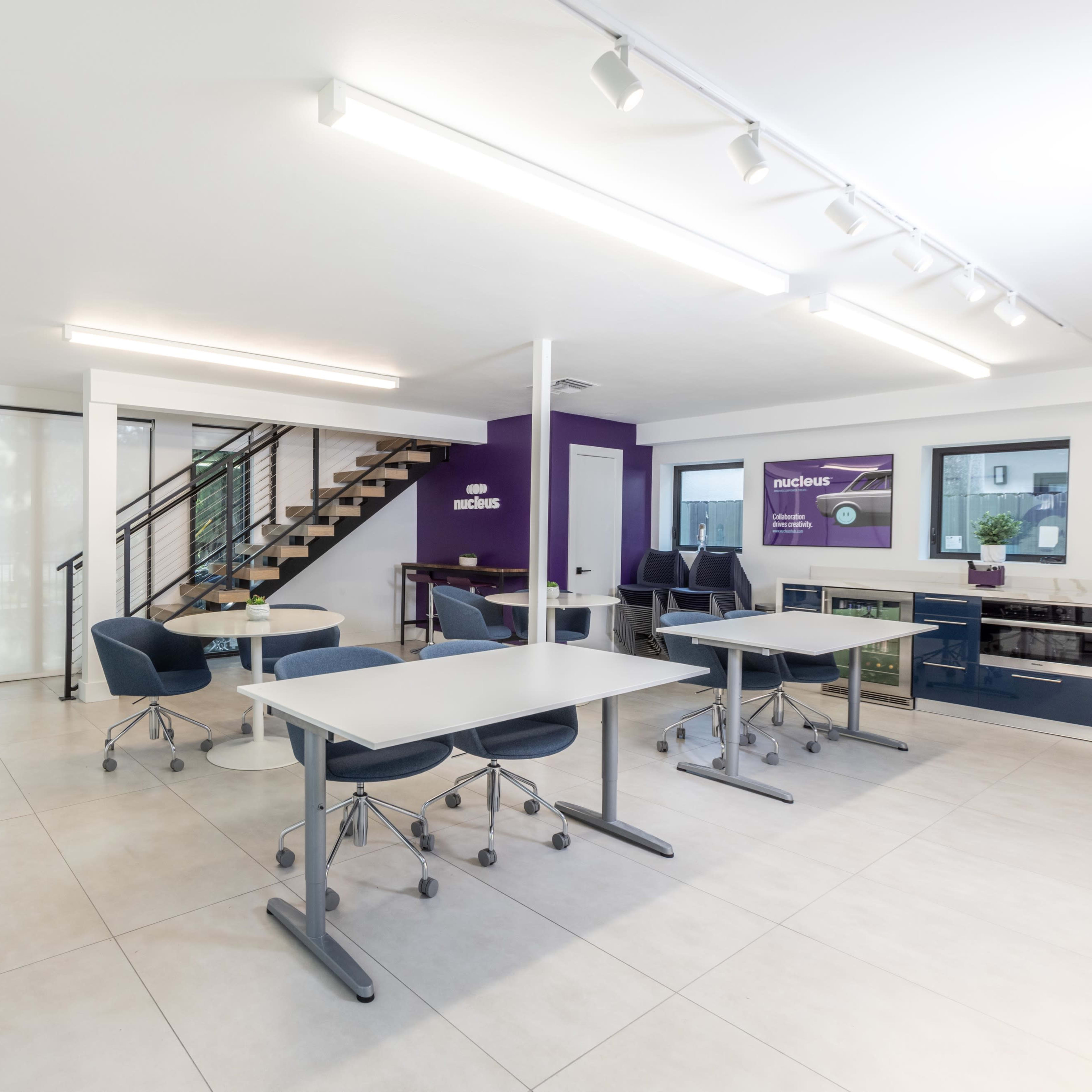 A modern office space featuring white walls, several tables, blue chairs, and a staircase, with a purple accent wall.