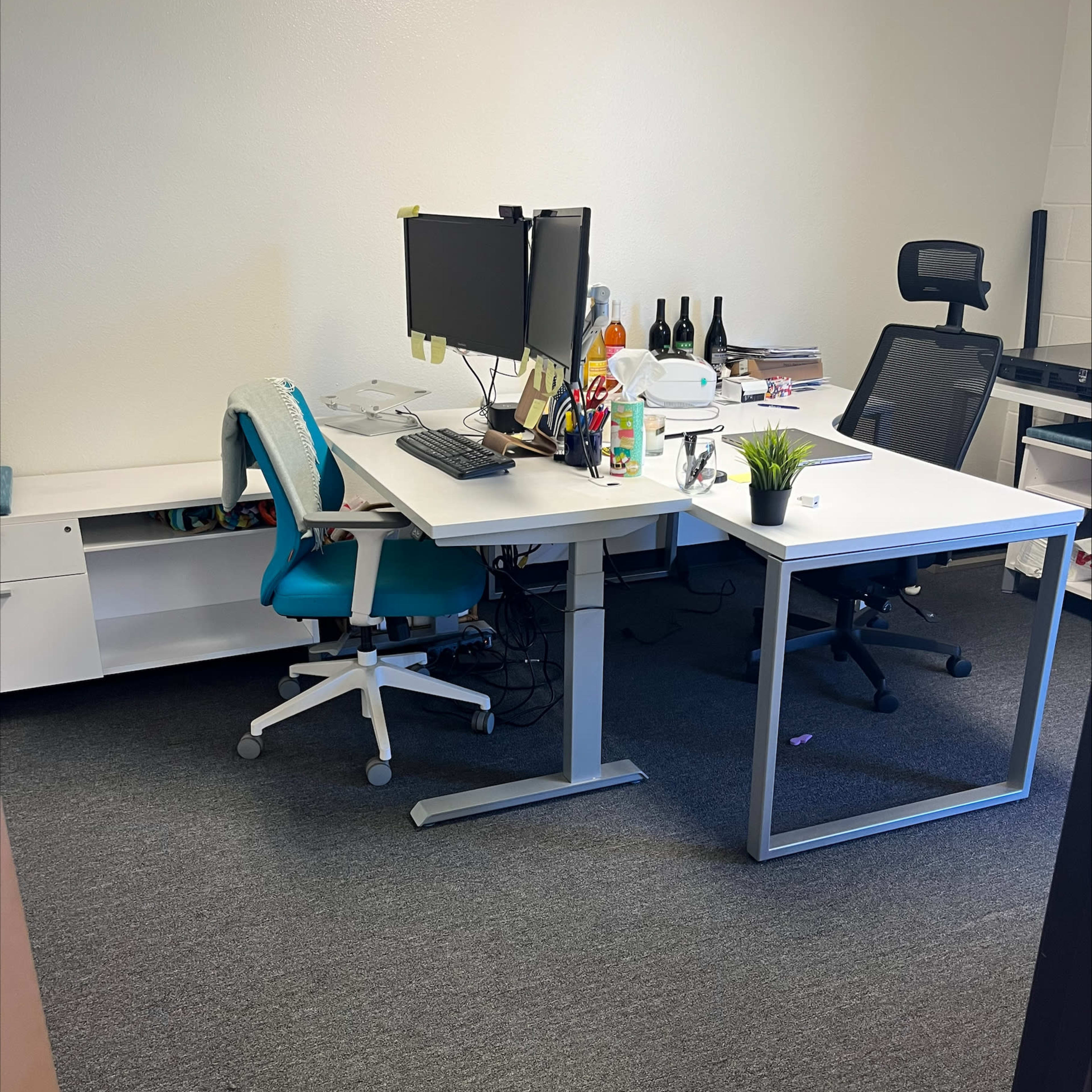 An office space features two desks, one with a computer monitor and office supplies, and the other equipped with a chair, set against a plain wall.