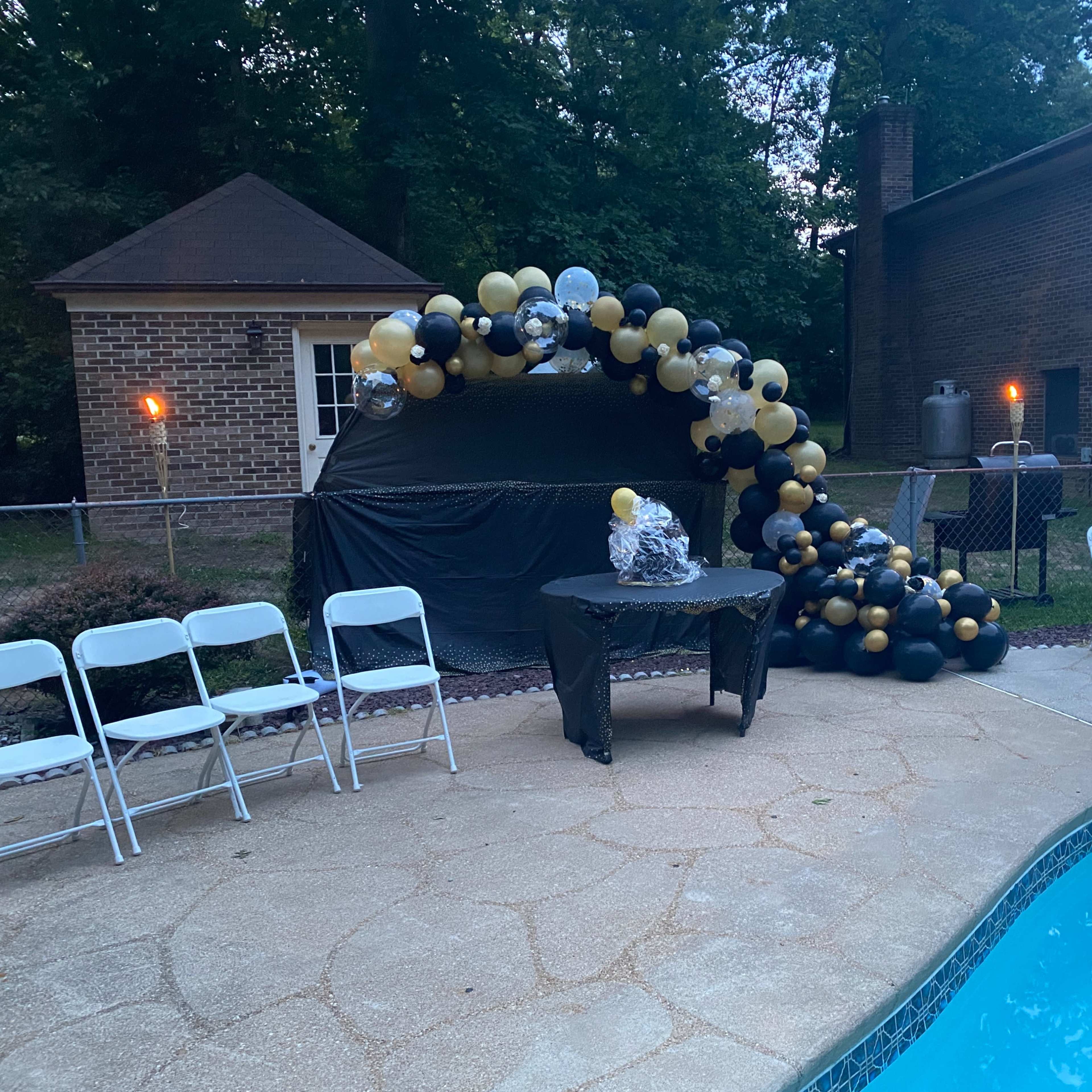 A decorated seating area with a table is set up by a swimming pool, featuring a balloon arch in black and gold colors nearby.