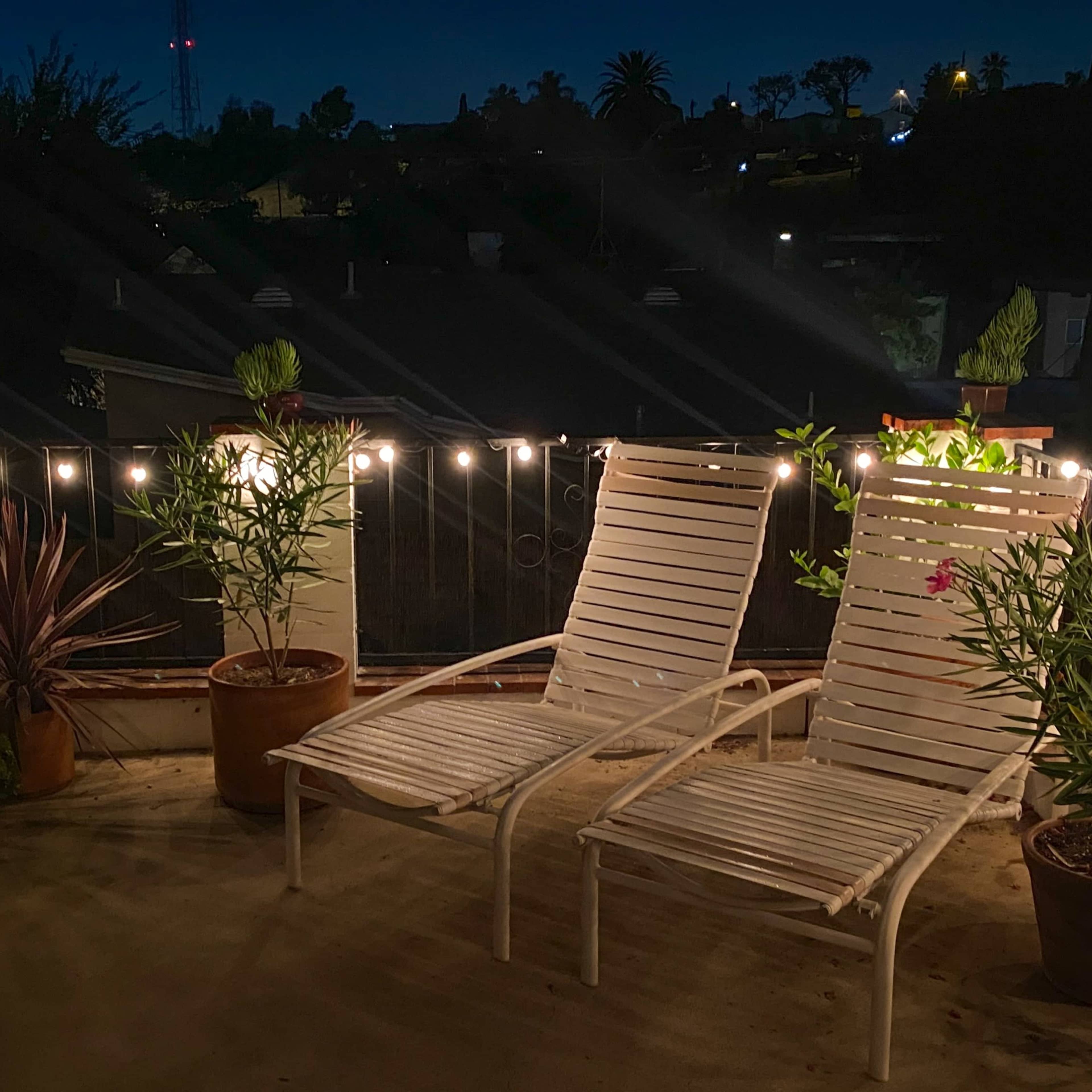 Two white lounge chairs are positioned on a terrace surrounded by potted plants and string lights against a night sky.