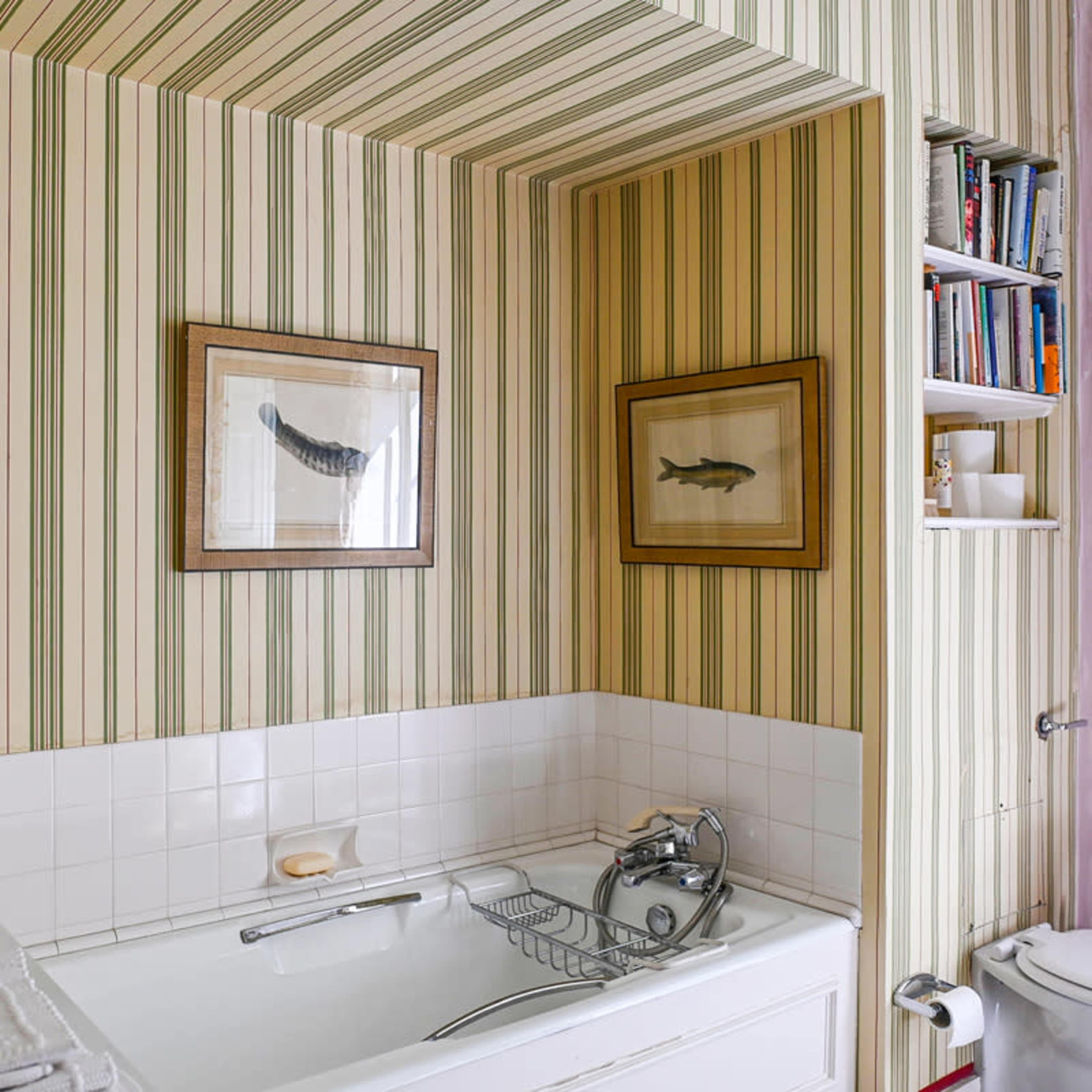 A bathroom with a tub surrounded by striped wallpaper, displaying framed fish prints and shelves containing toiletries and books.