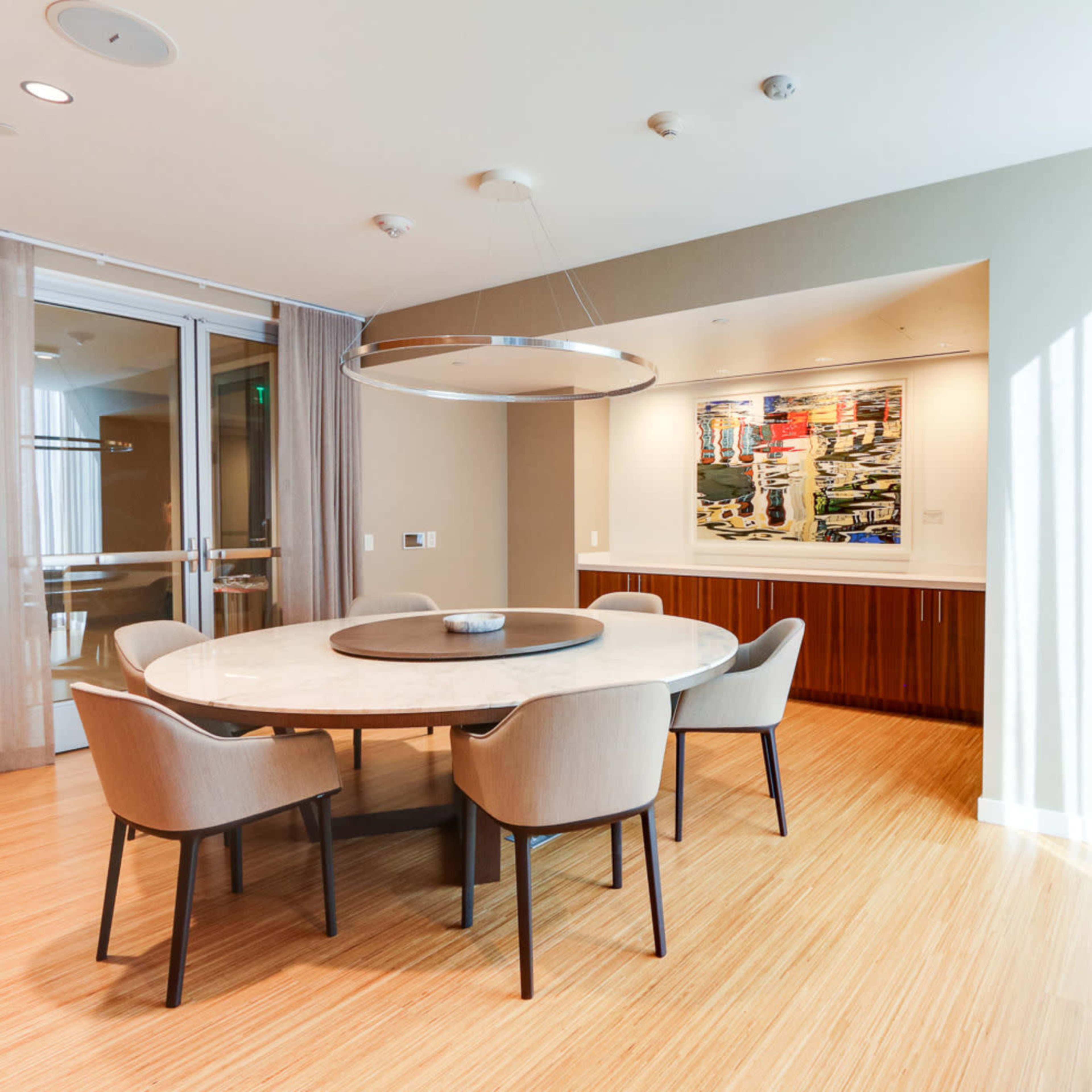 A round dining table with light-colored chairs is set in a brightly lit room featuring a large artwork on the wall.