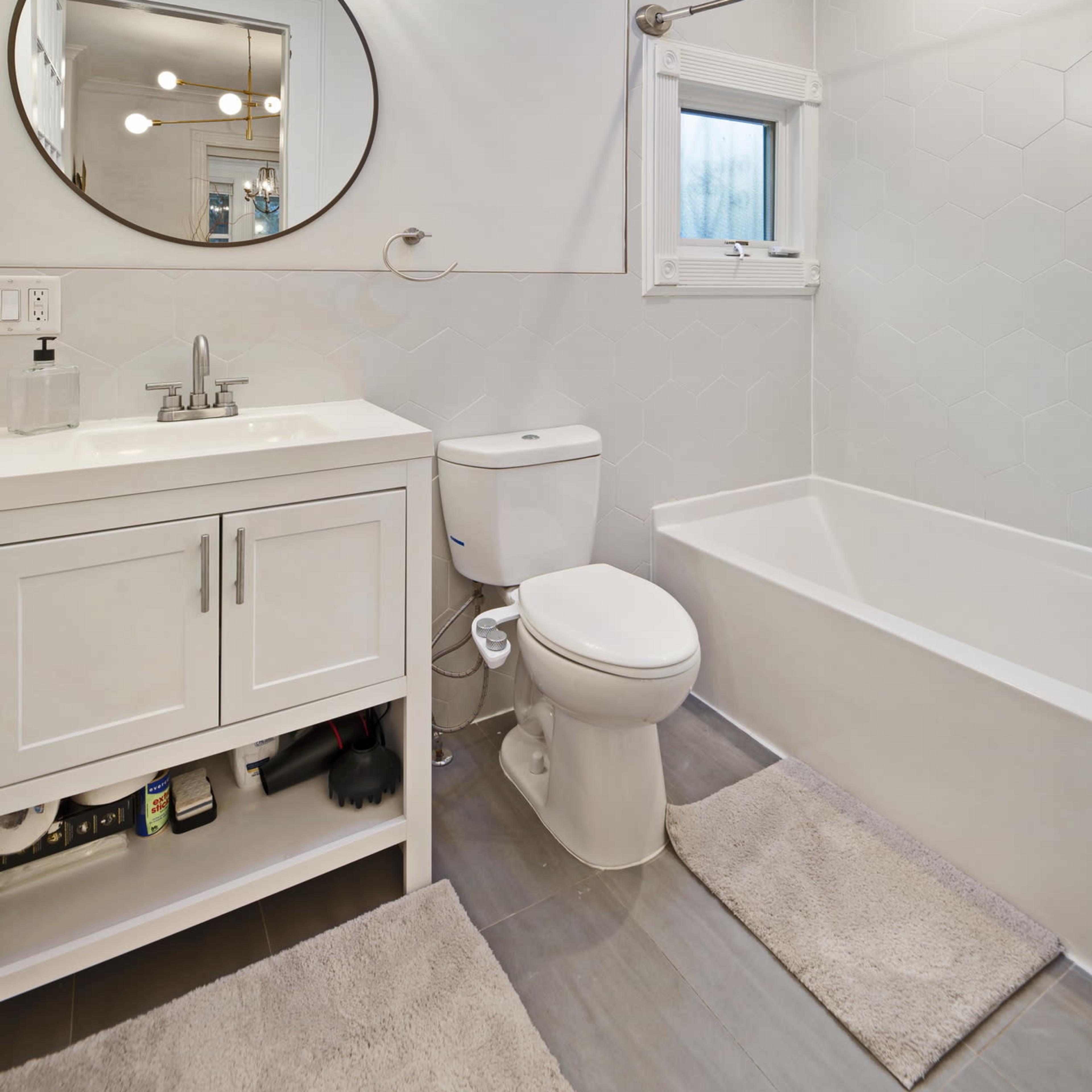 A modern bathroom featuring a white vanity with a round mirror, a bathtub, a toilet, and gray tile flooring.