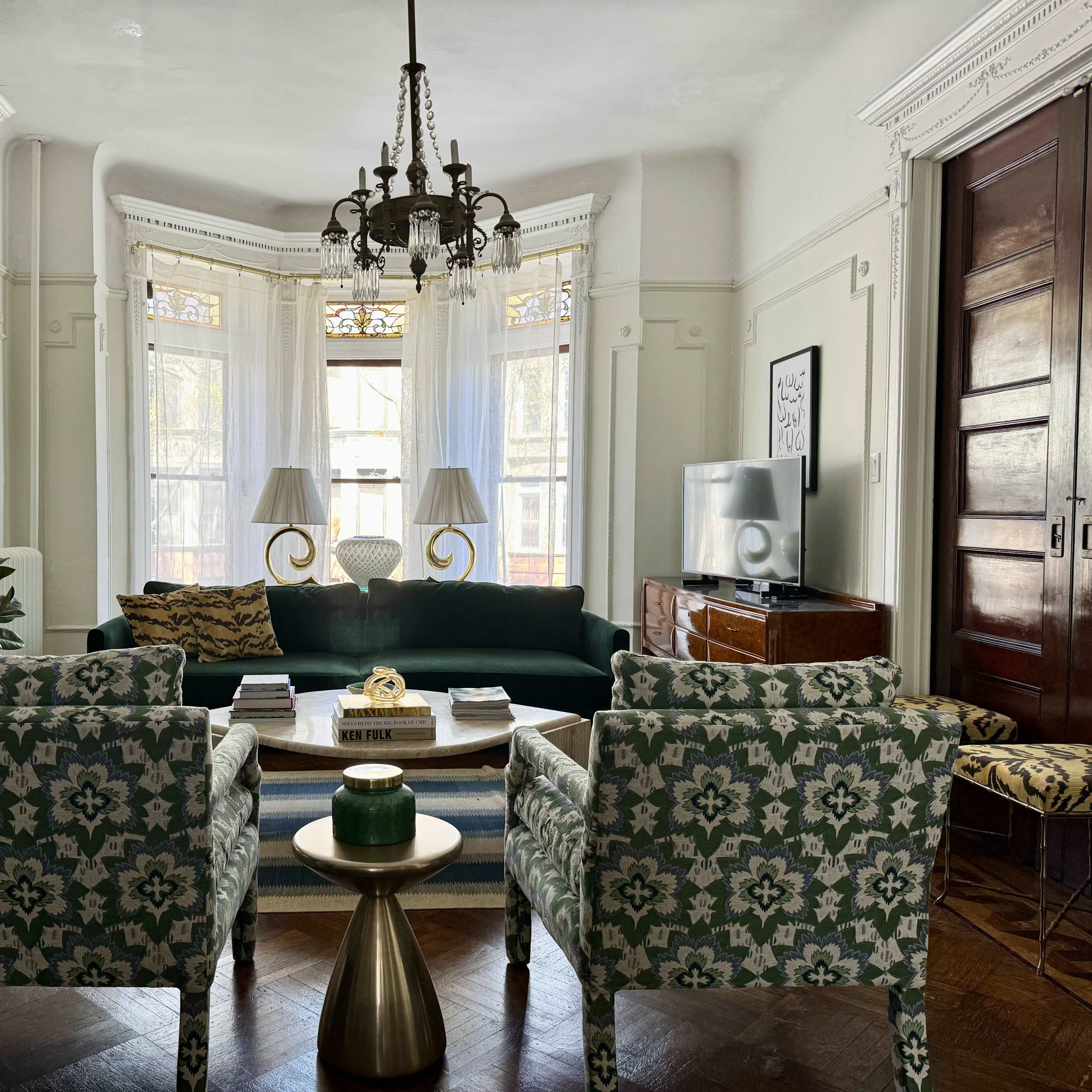 A brightly lit living room with a green sofa, two patterned armchairs, a coffee table, and large windows adorned with white drapes.