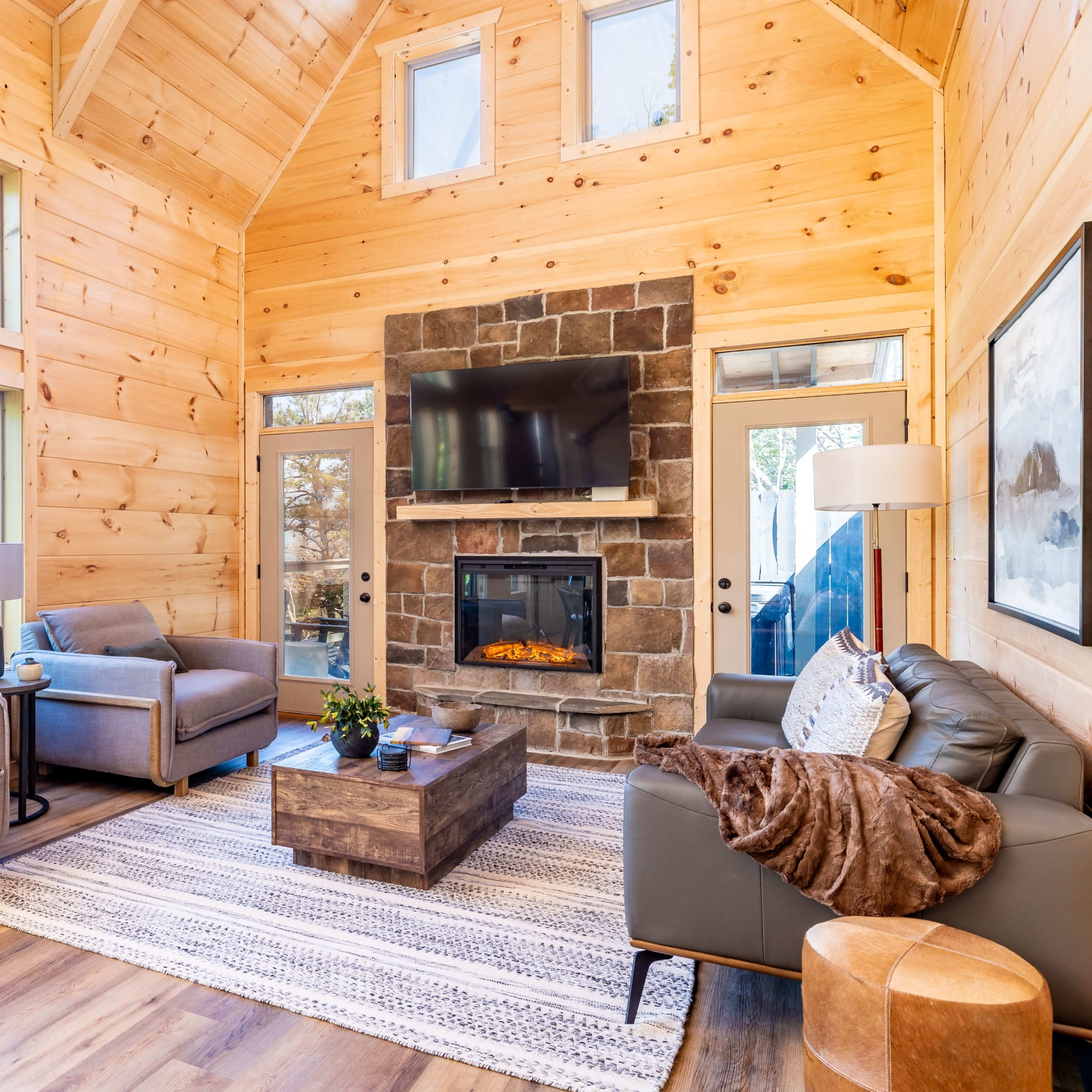 A modern living room features large windows, wooden walls, comfortable seating, a stone fireplace, and a coffee table on a textured rug.