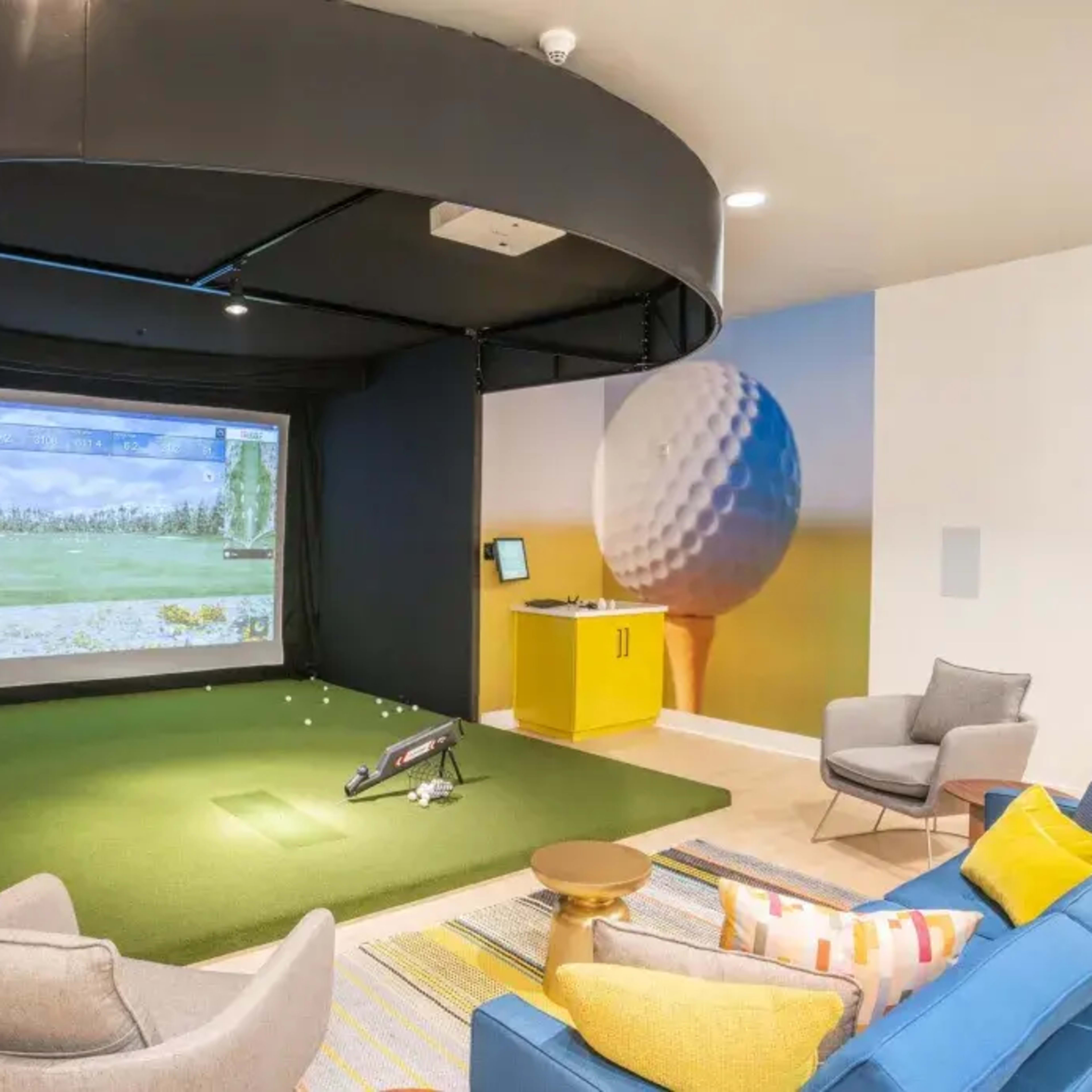 An indoor golf simulation room featuring a large screen, synthetic grass, and modern seating arrangements.