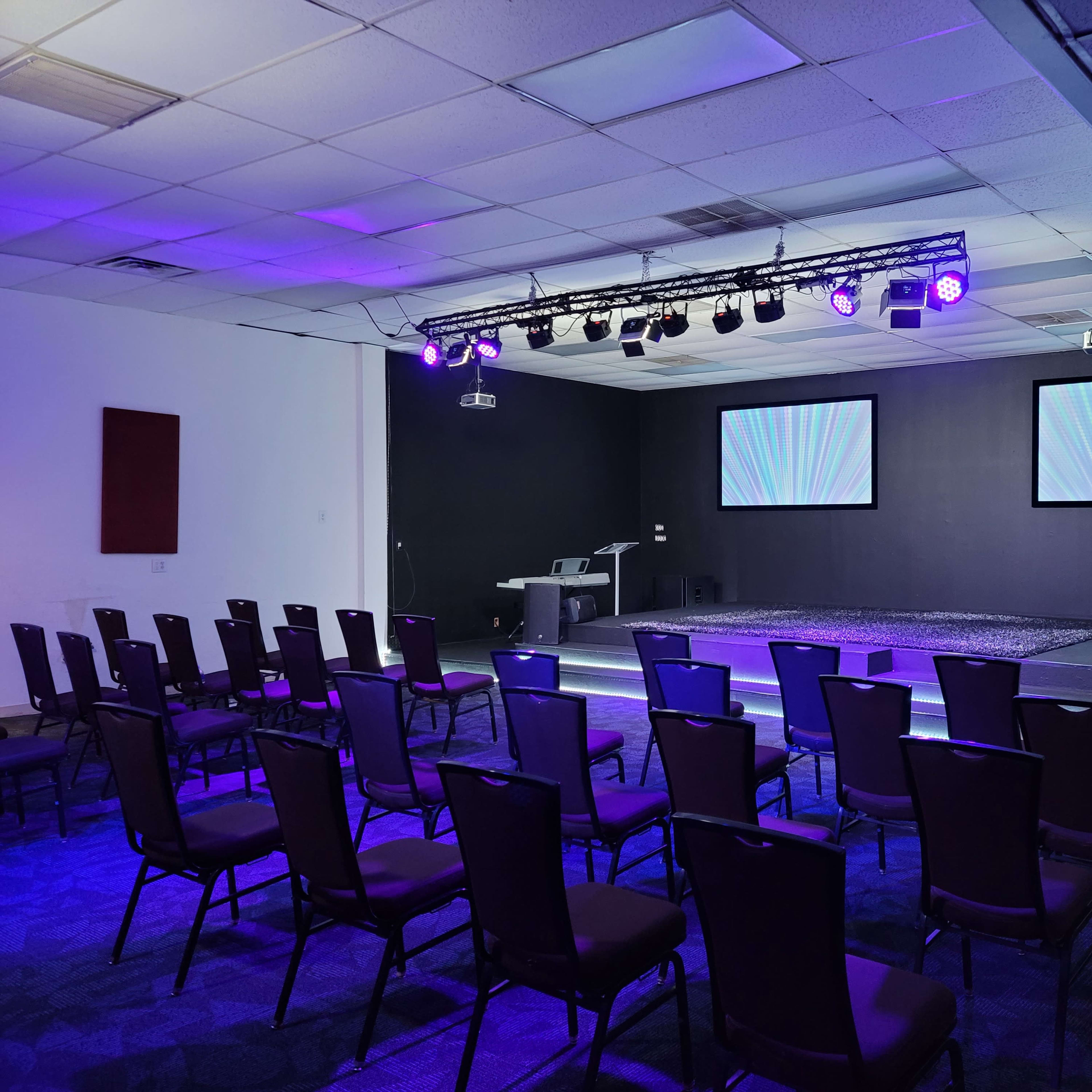 A prepared indoor event space with rows of chairs facing a stage that has two projection screens and stage lighting.