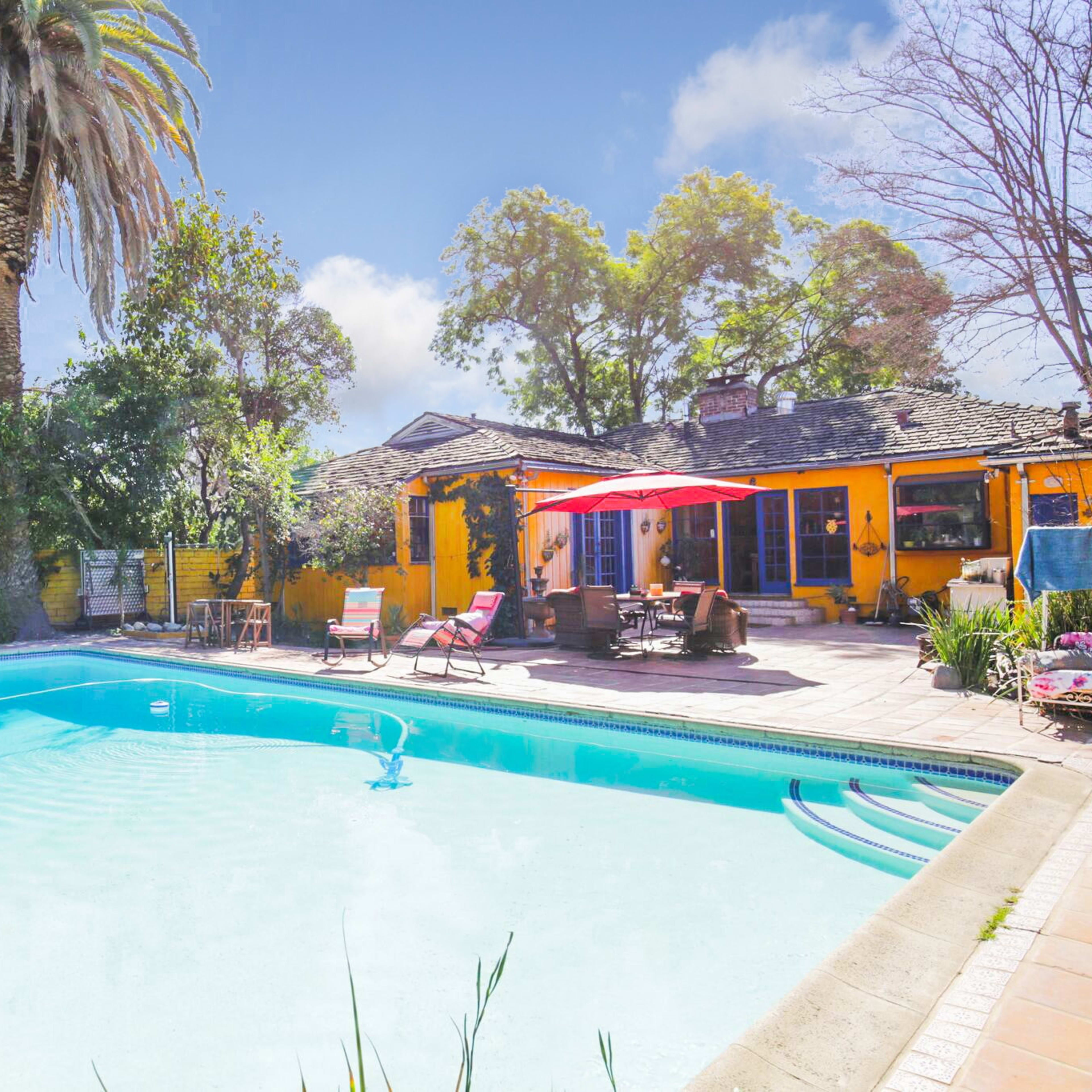 A backyard with a swimming pool surrounded by palm trees and lounge chairs, featuring a house with a patio and colorful walls.