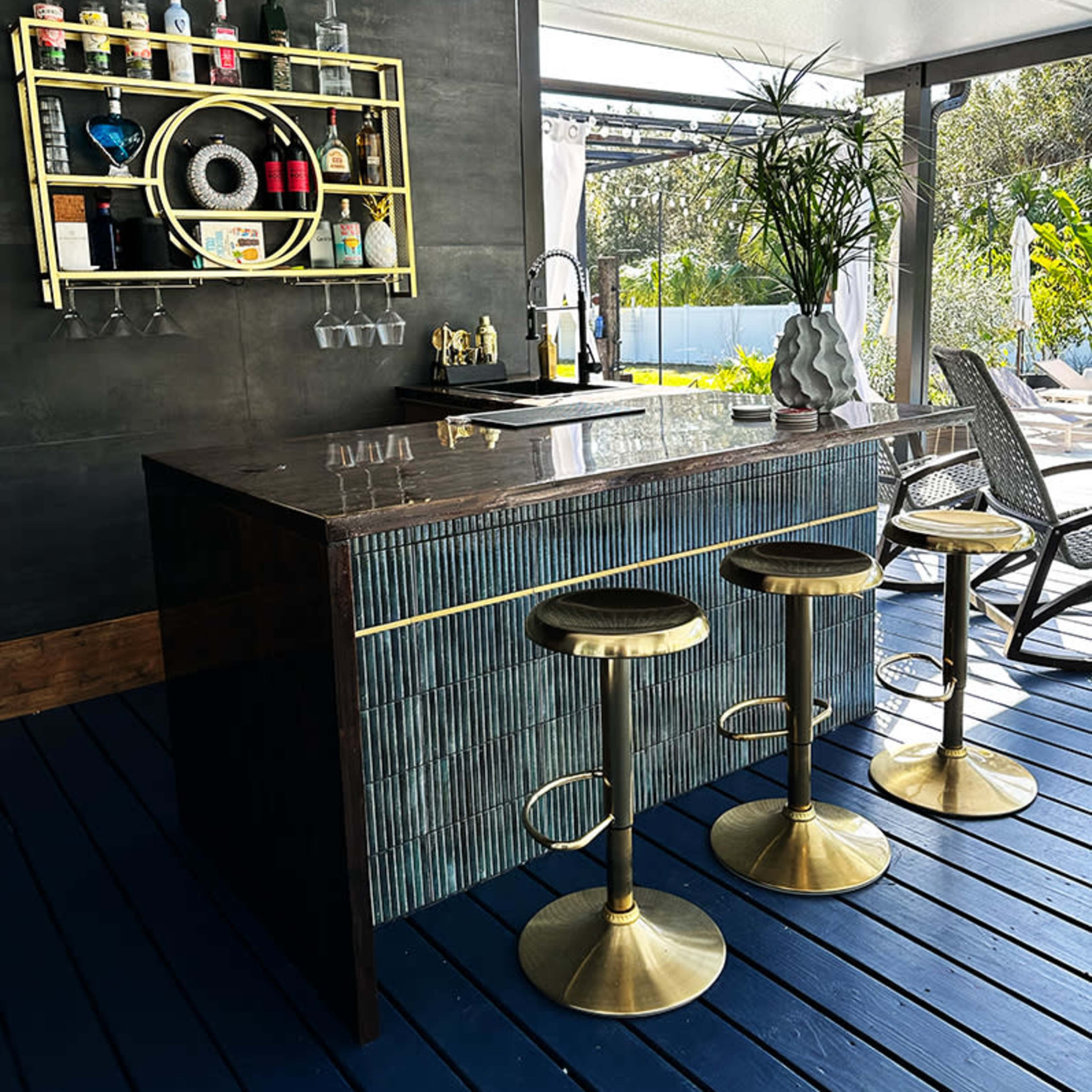 A modern outdoor bar with three brass stools, a decorative backlit shelf holding various bottles, and a sleek countertop.