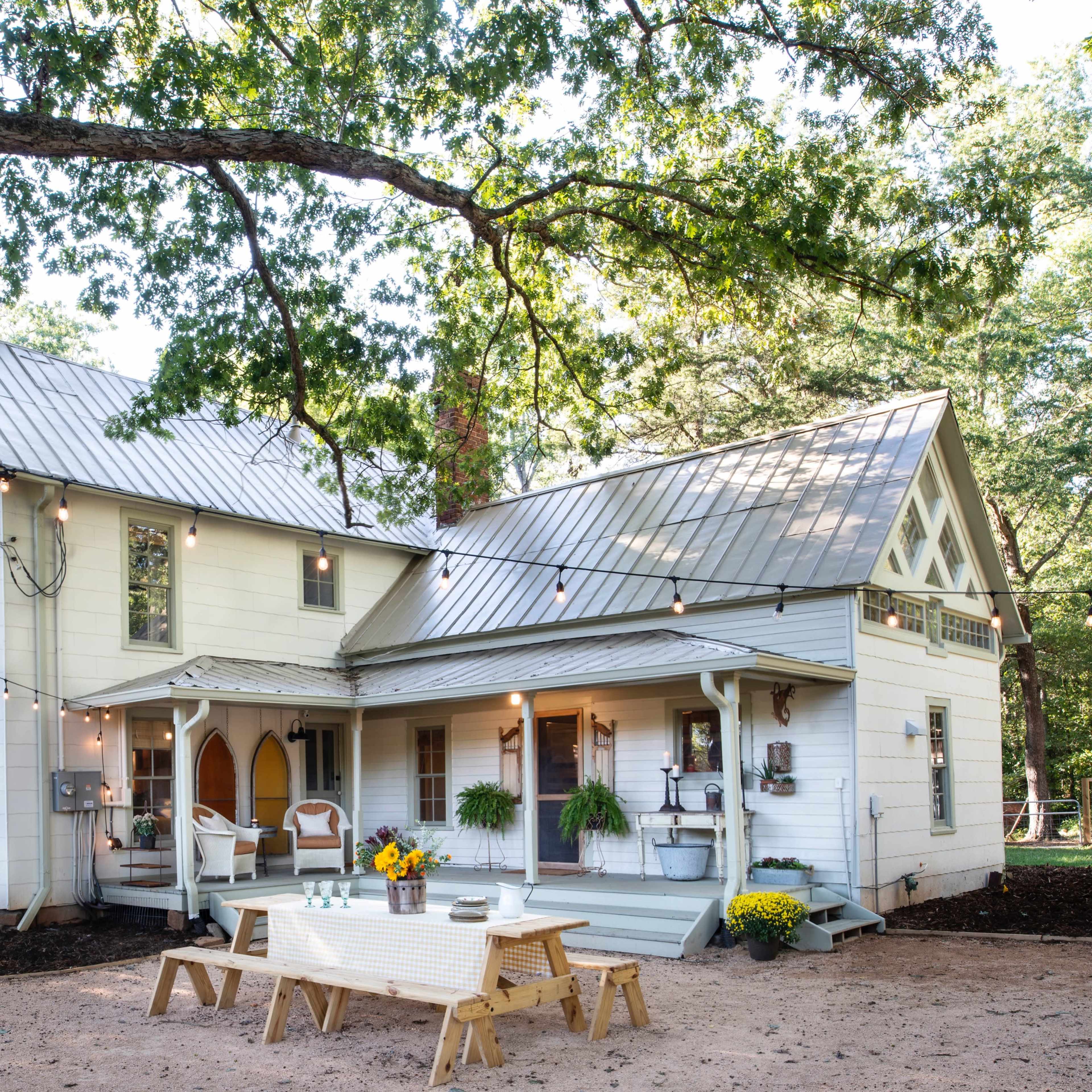 A two-story white farmhouse with a metal roof and a patio area is surrounded by trees and string lights.