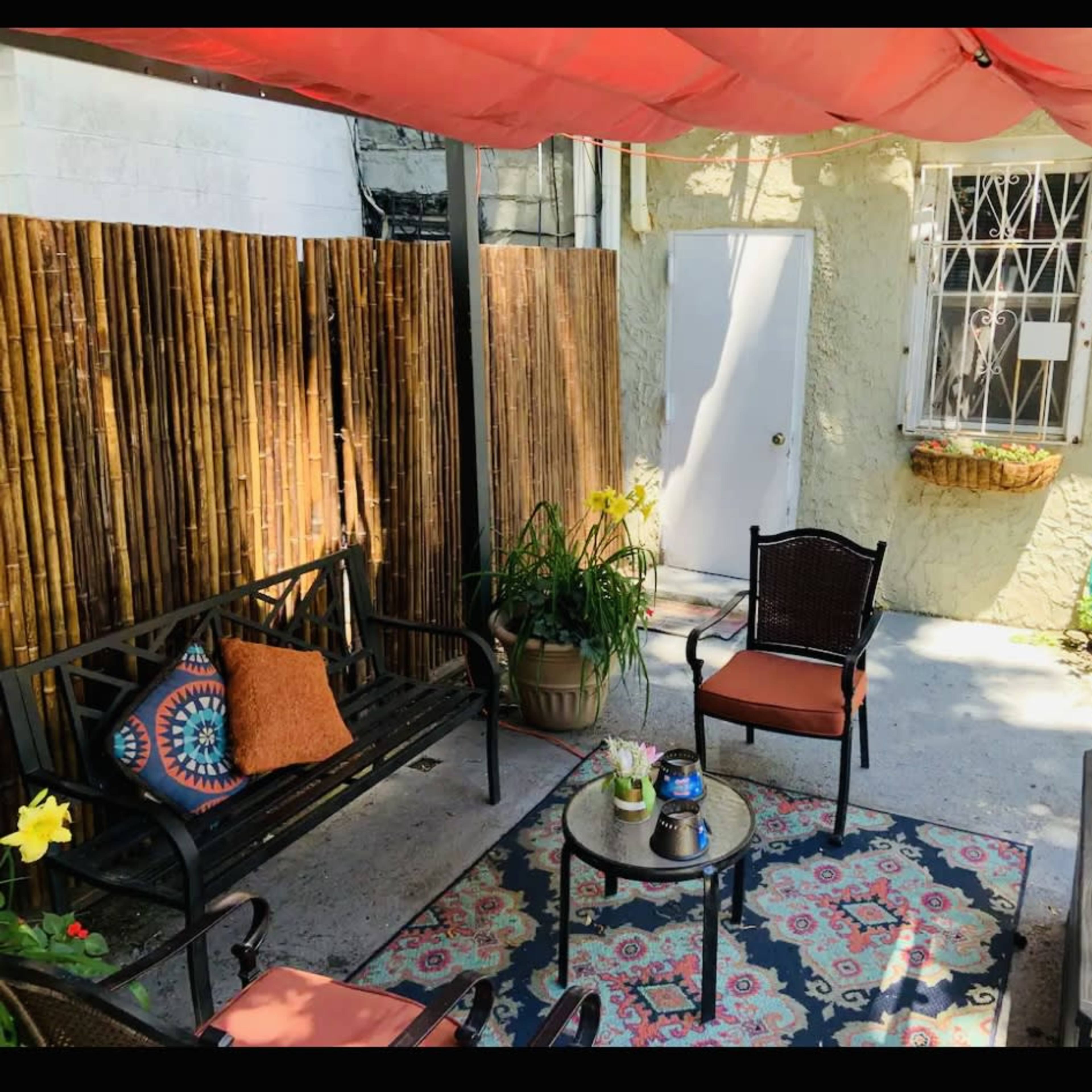 A patio features a table with chairs, a decorative rug, potted plants, and a shaded area with a canopy.