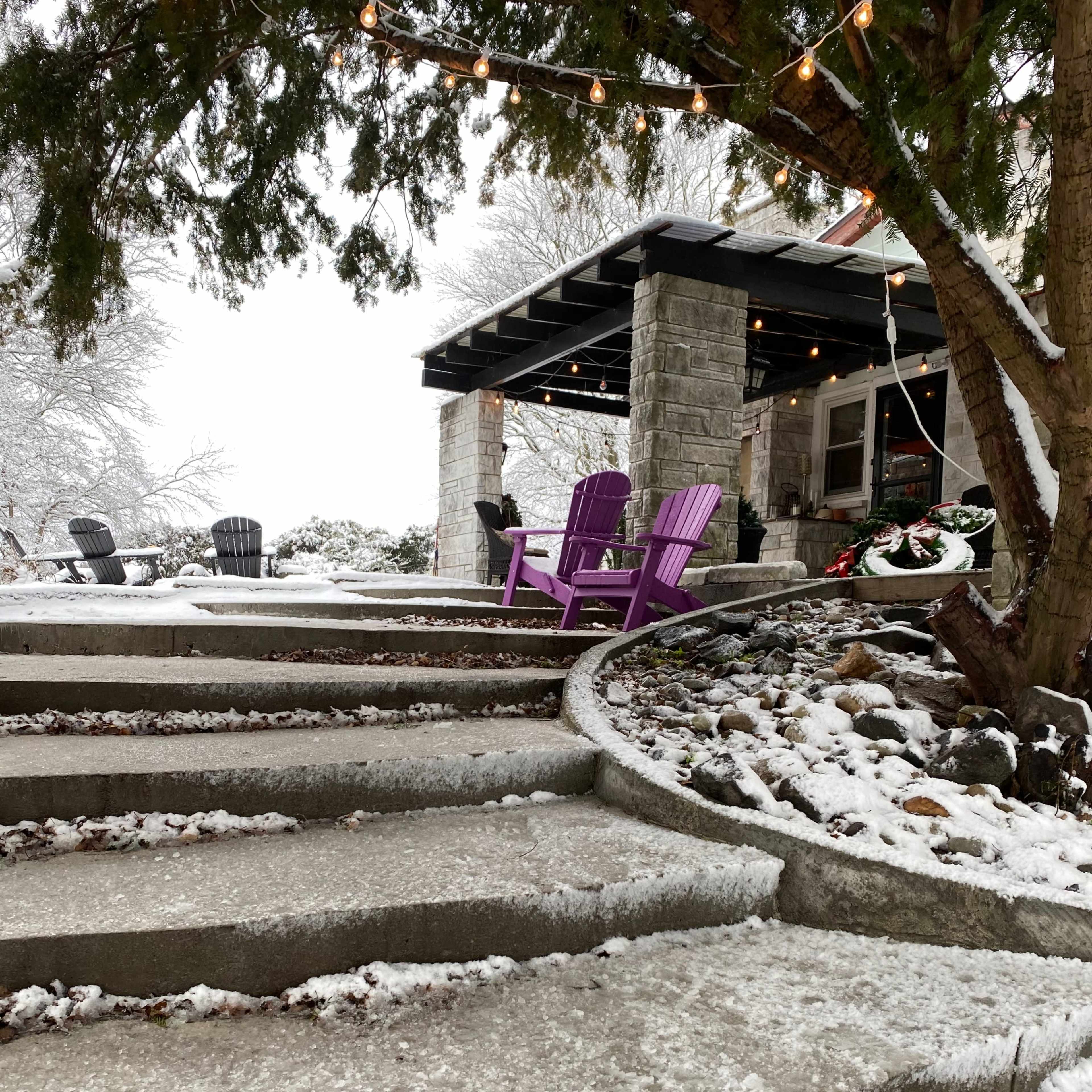 A winding stone pathway leads to a porch with purple chairs, surrounded by snow and decorative outdoor lighting.