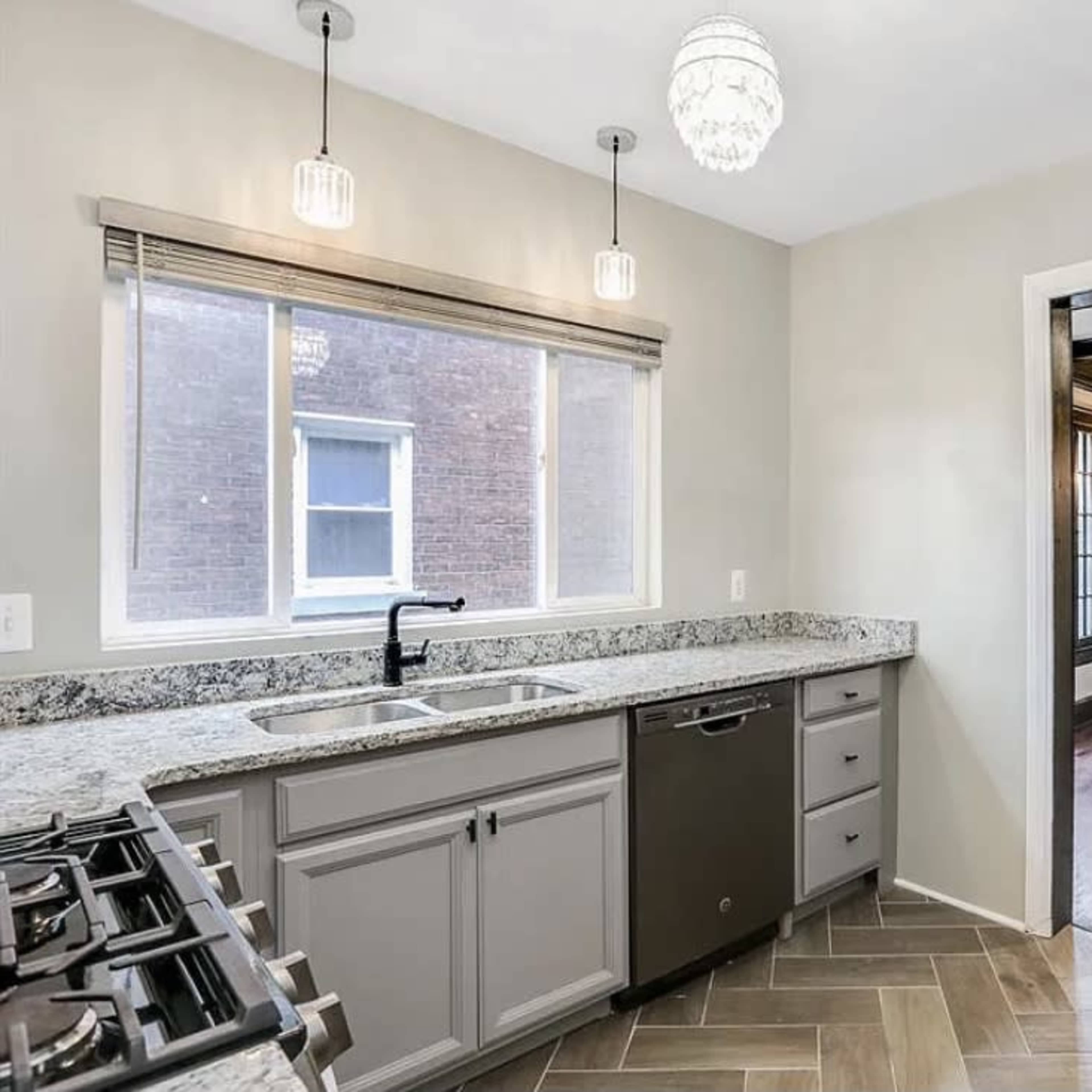 A modern kitchen area with granite countertops, a double sink, a dishwasher, and a view into a living room featuring wooden beams and a chandelier.