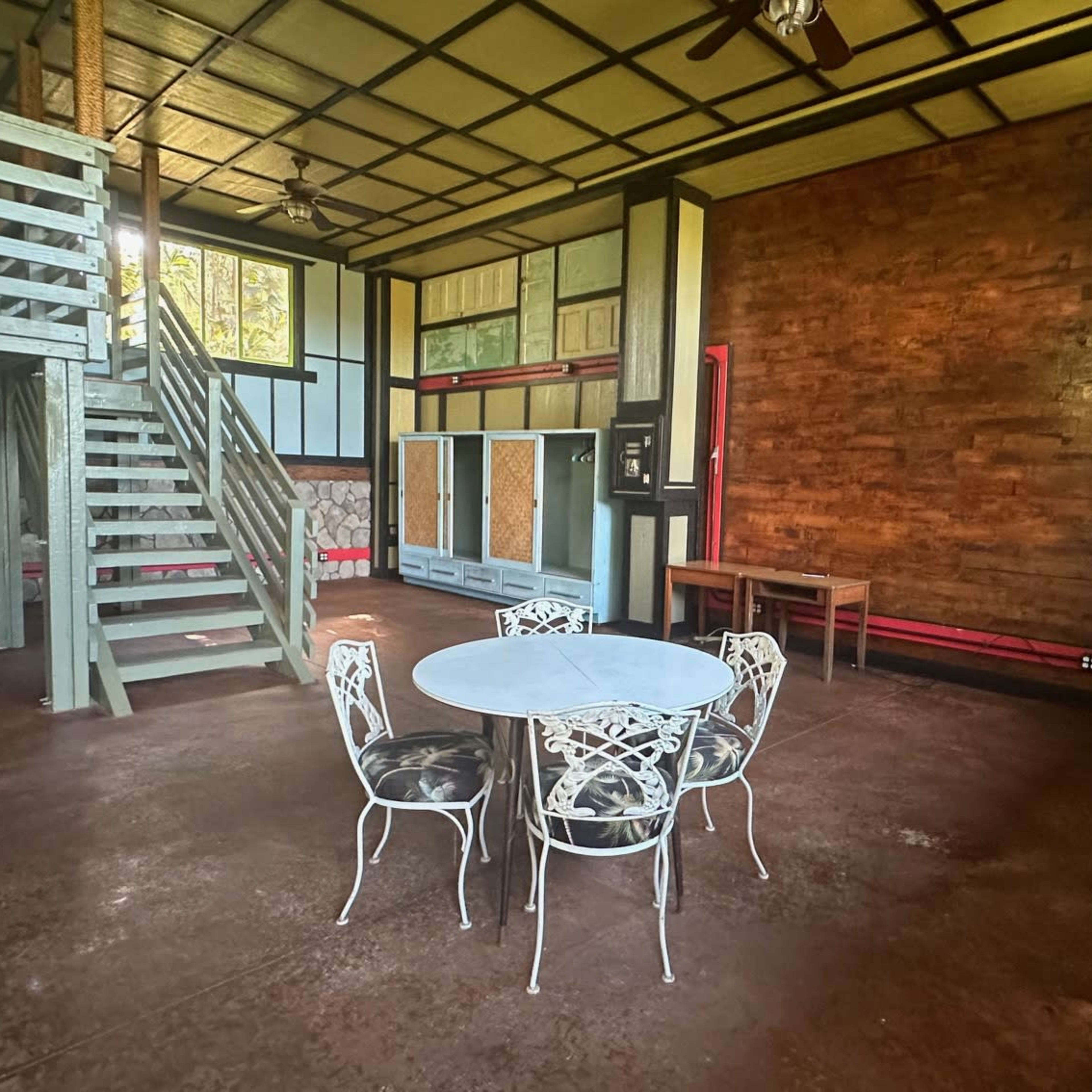 A spacious room features a round dining table with white chairs, a staircase in the background, and a wooden wall accent.
