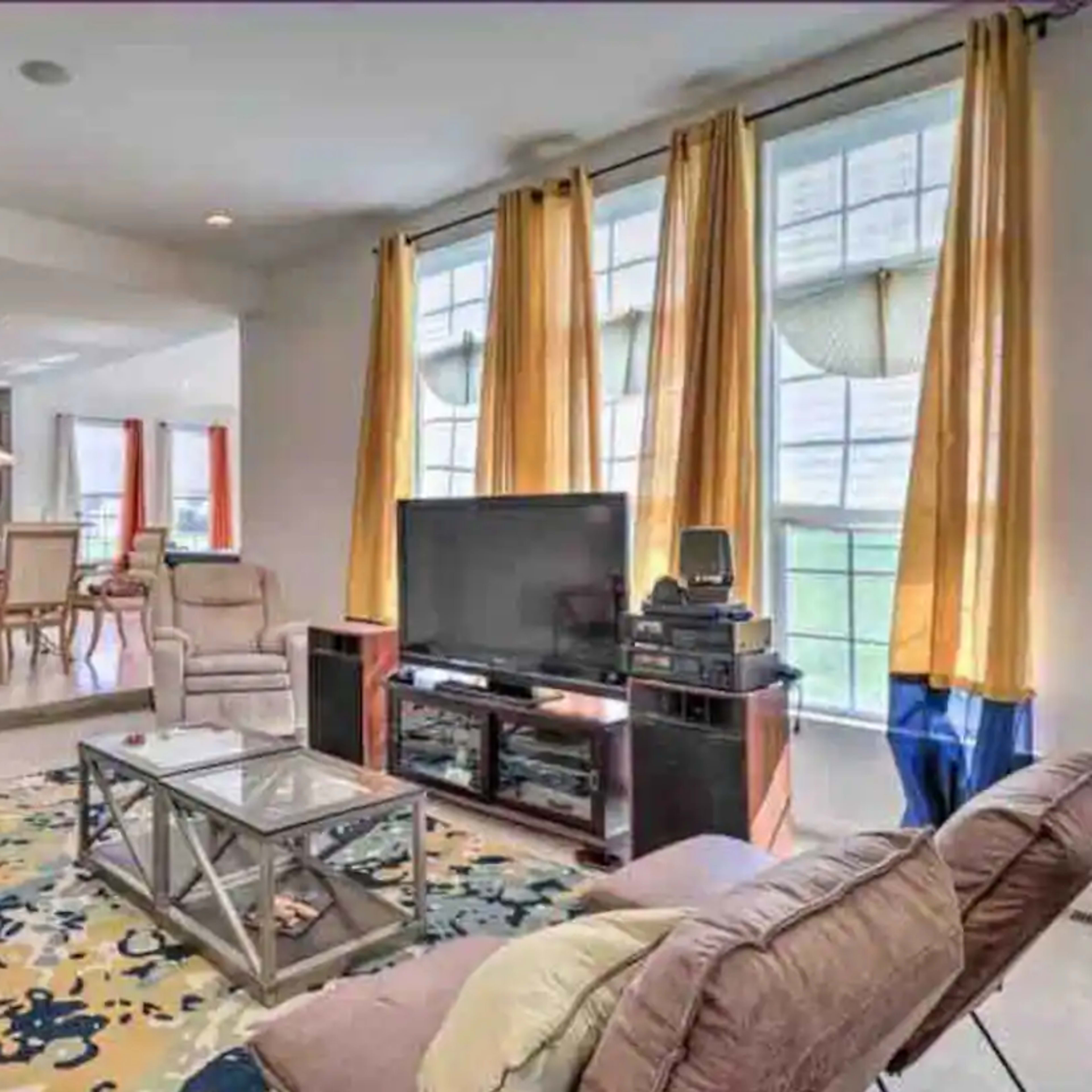 A living room with a large window dressed in yellow curtains, a television on a stand, and a coffee table on a colorful rug, leading into a dining area in the background.