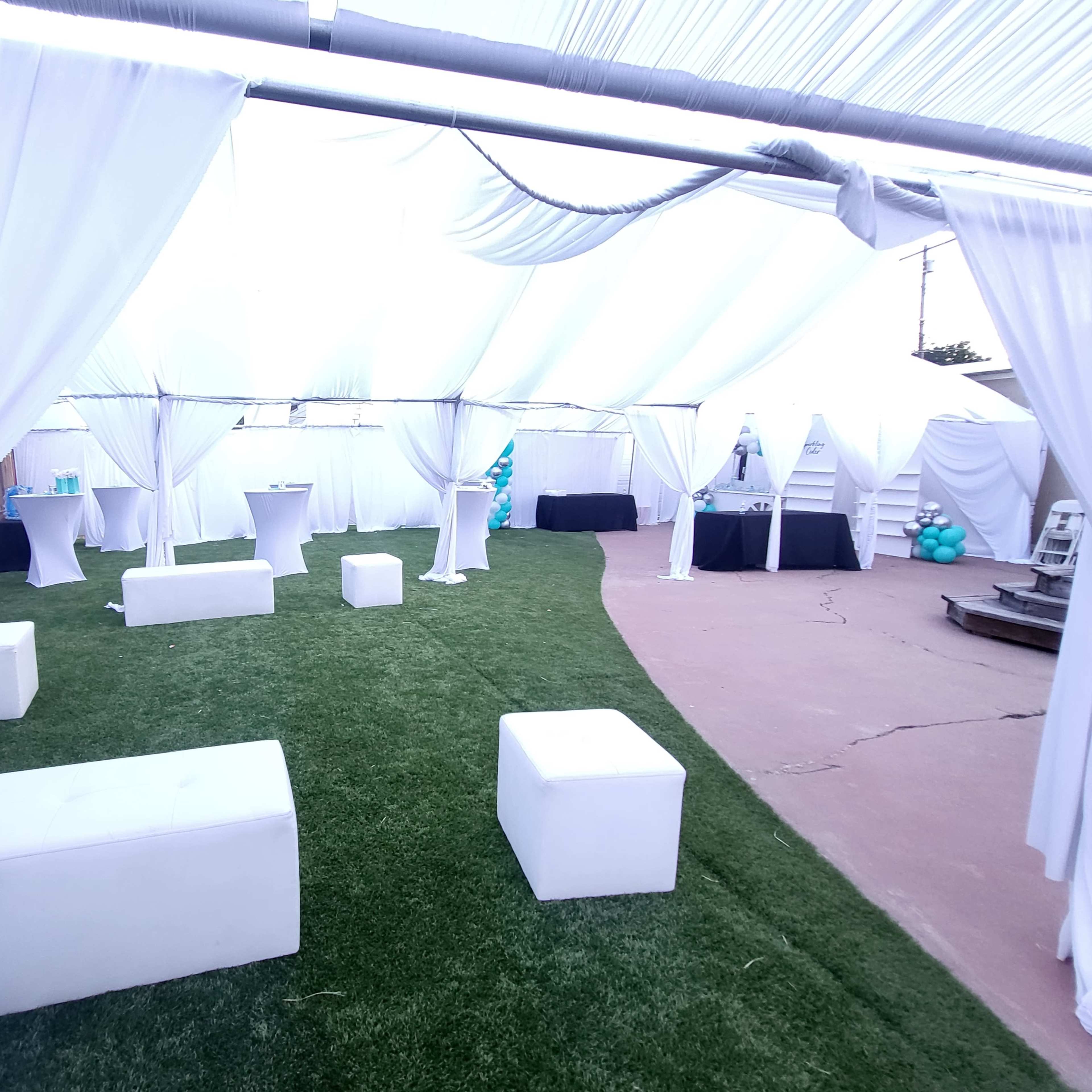 A tented outdoor event space featuring white draped fabric, green artificial grass, and various seating arrangements with cocktail tables and a black display table in the background.