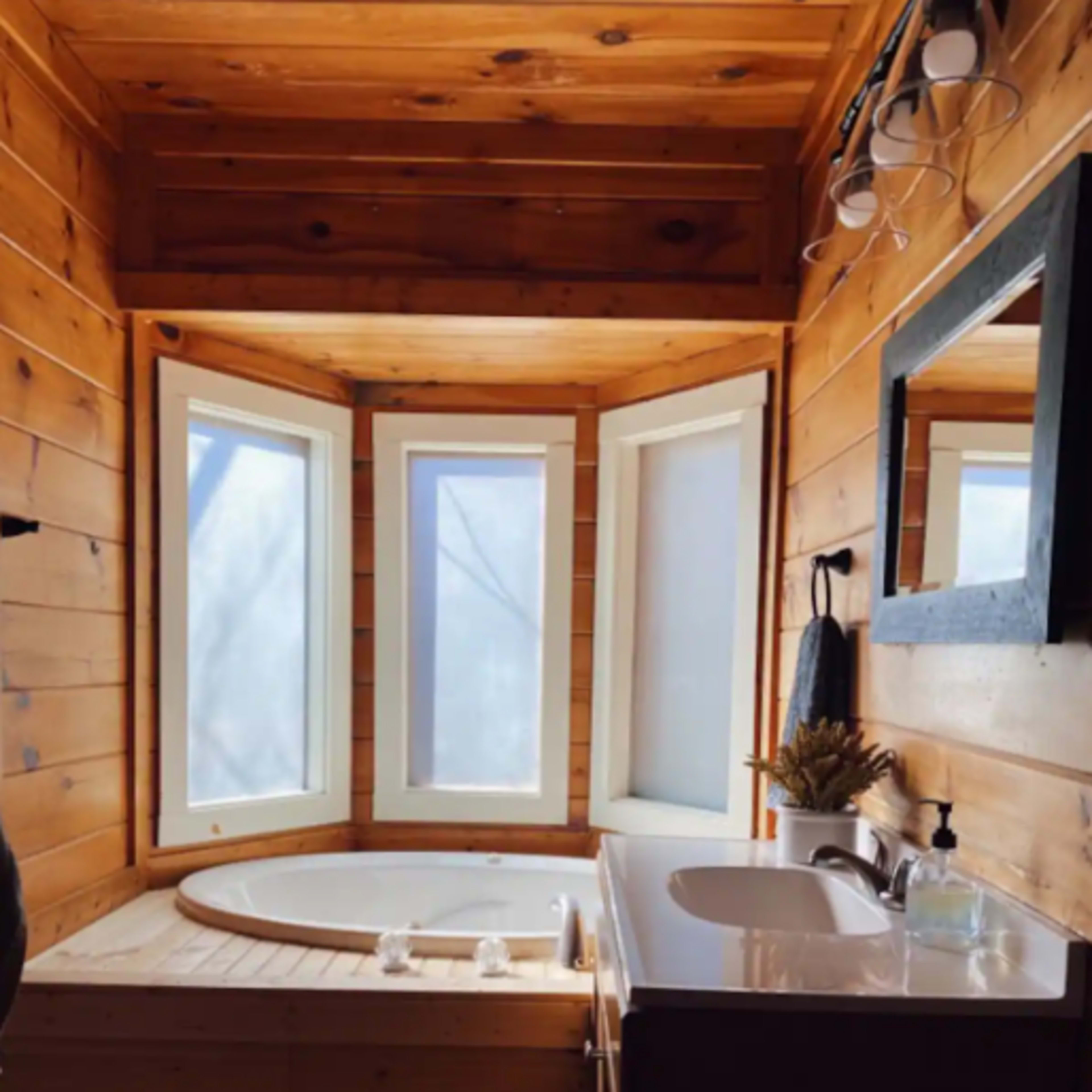 A wooden bathroom featuring a corner bathtub with three large windows and a sink with a mirror above it.