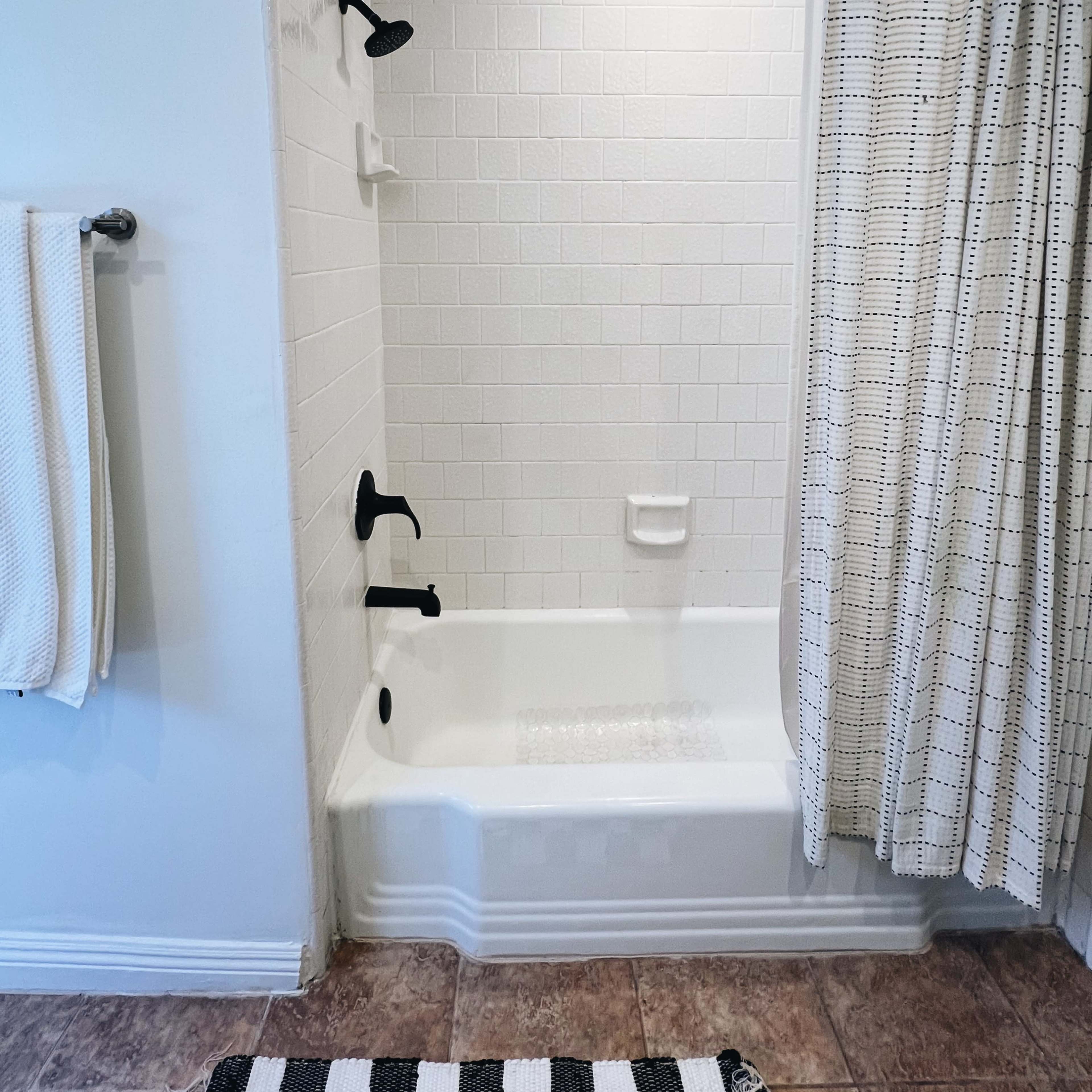A bathroom with a white tiled shower area, a bathtub with a black shower fixture, a patterned shower curtain, and a towel rack on the wall.