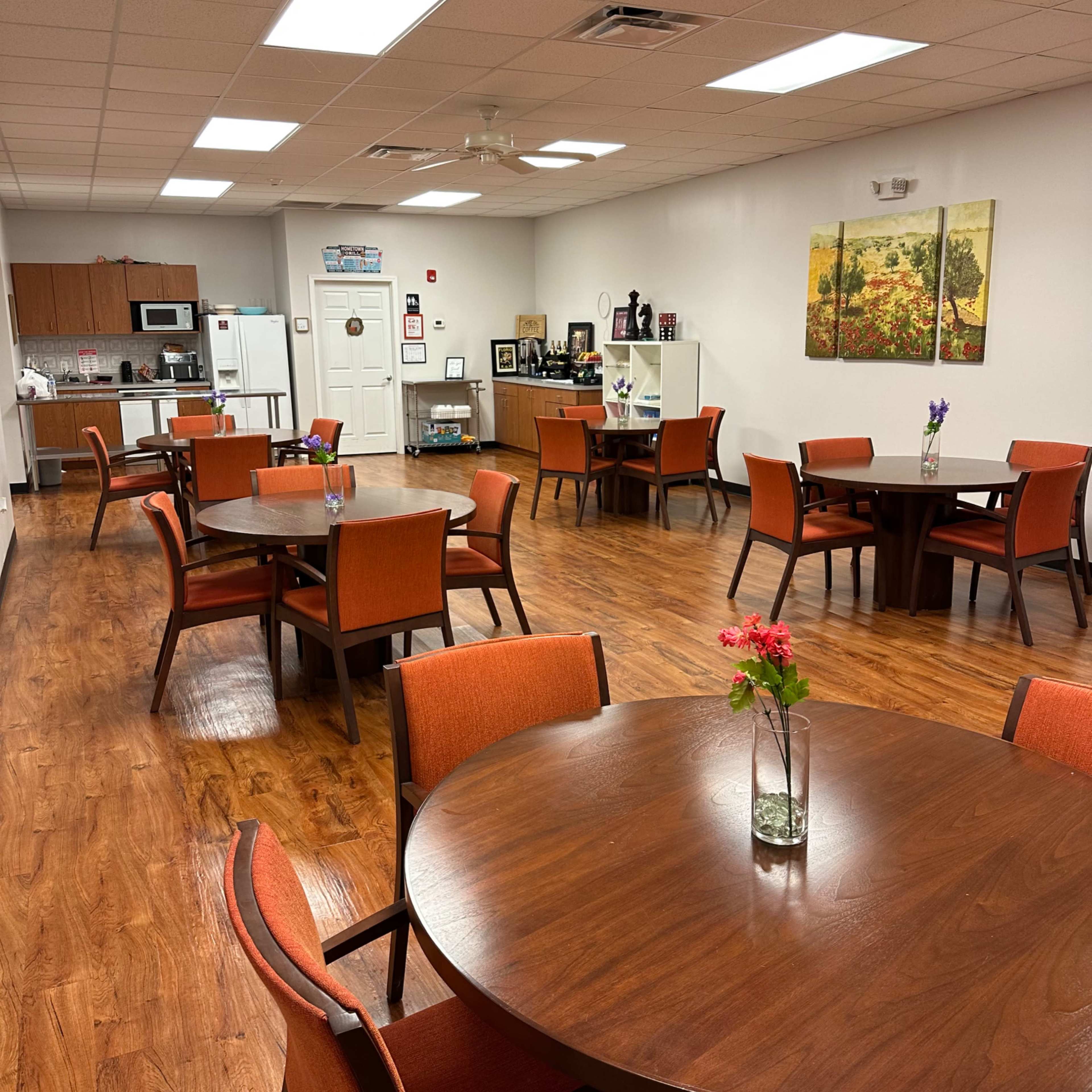 A dining area with wooden tables and orange chairs, equipped with a kitchenette in the background.