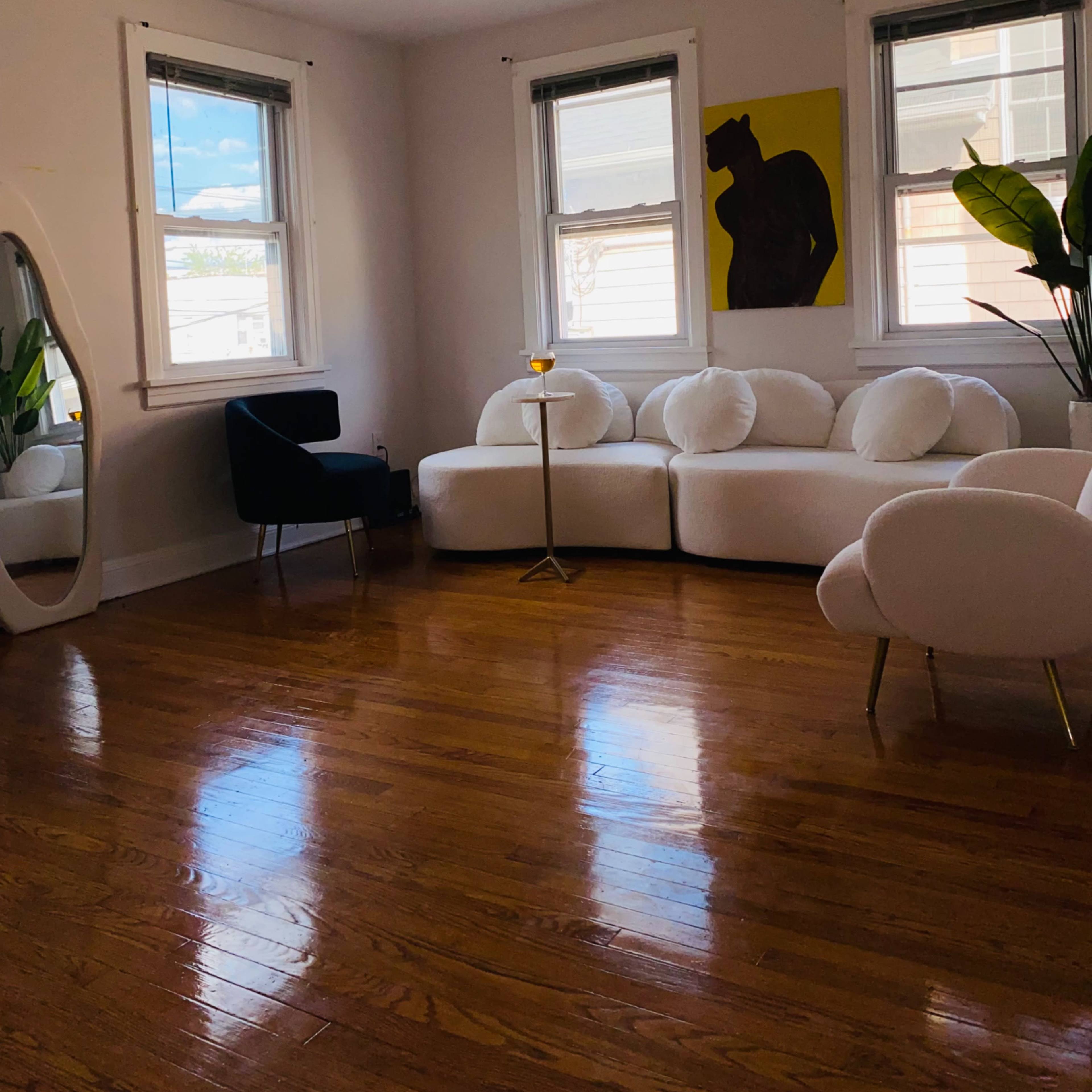 The room features white couches, a black chair, and a tall mirror, with light coming through two windows and a piece of artwork on the wall.