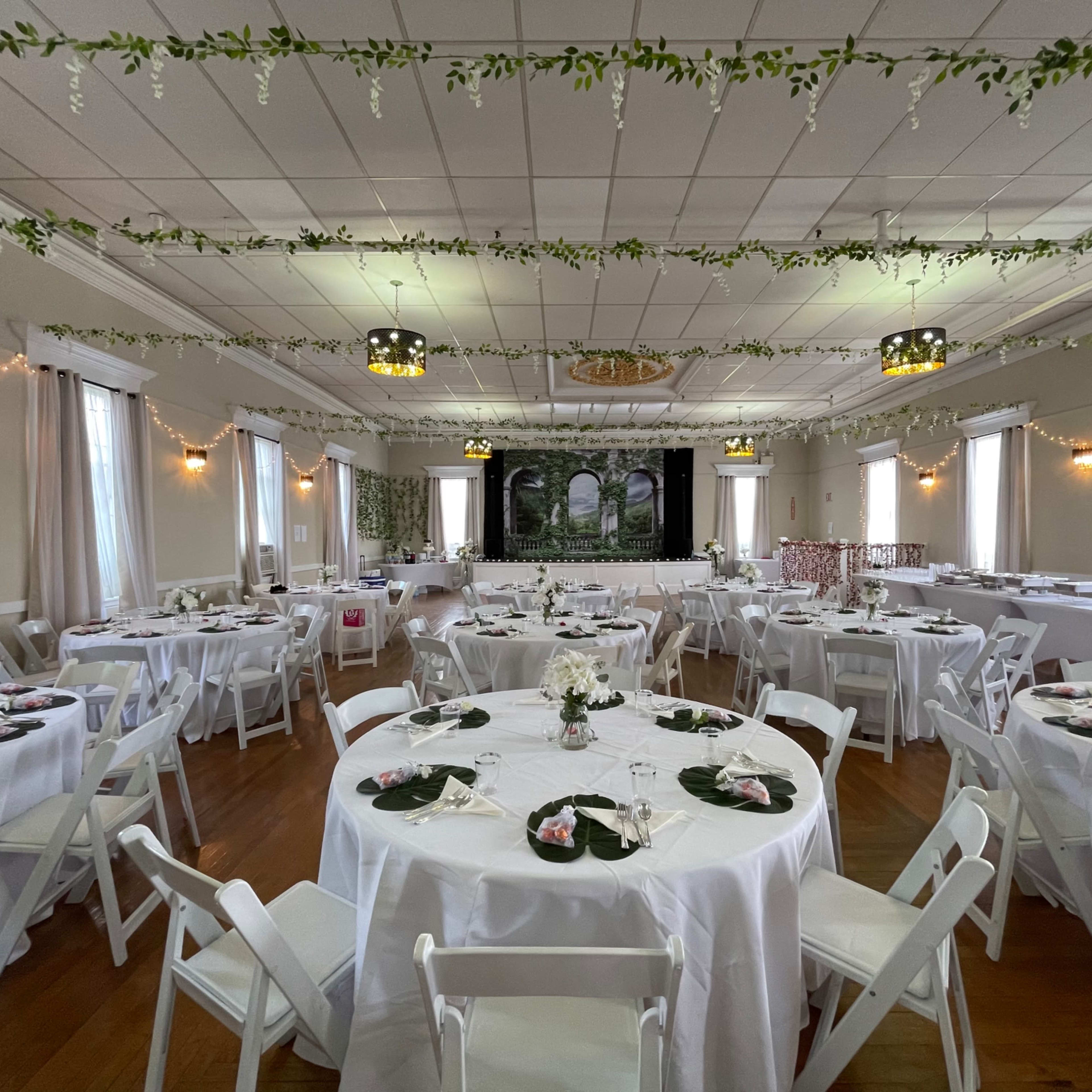 A banquet hall set up for an event, with round tables adorned with centerpieces and white chairs, all arranged under decorative greenery and lighting.