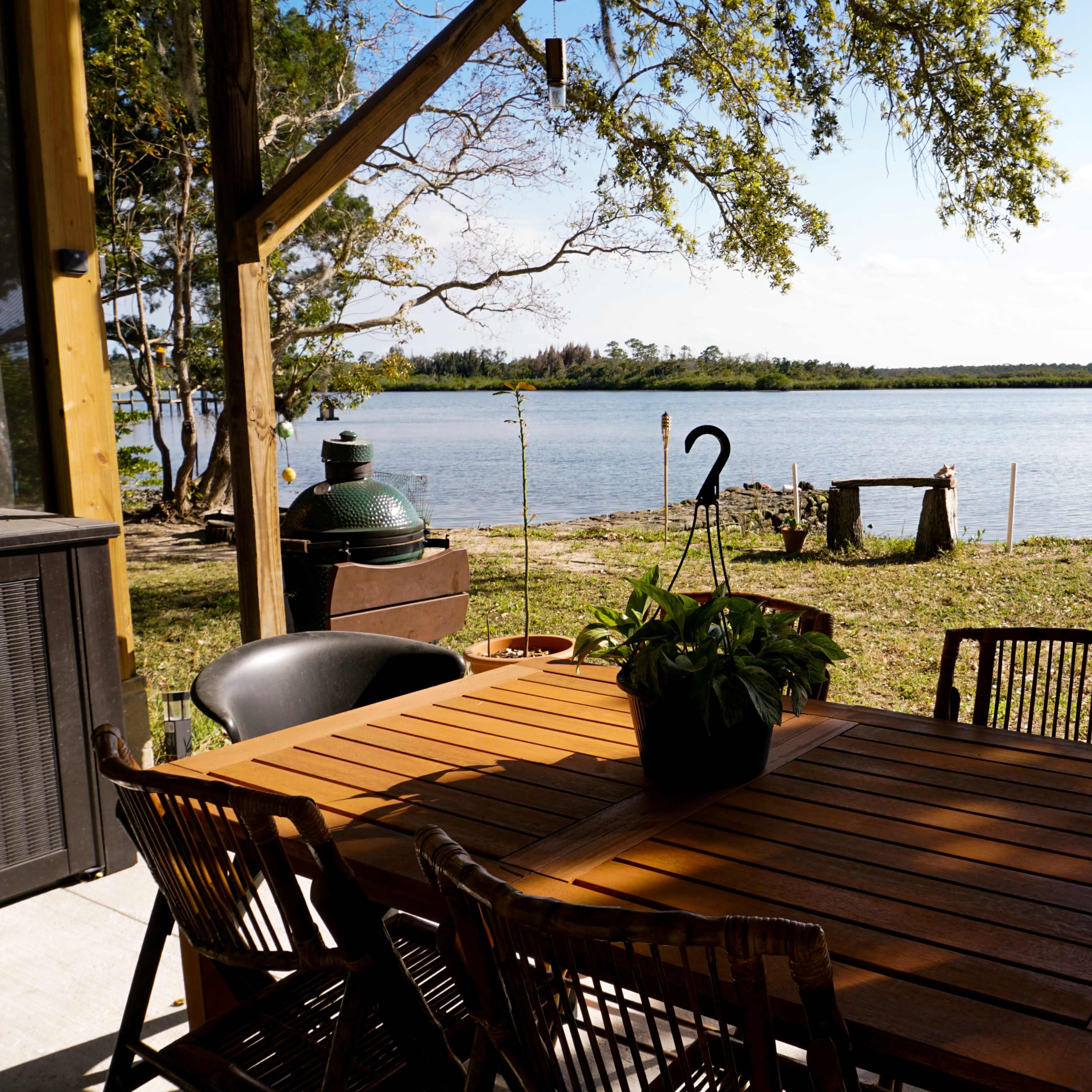 A wooden outdoor dining table with chairs overlooks a calm river and grassy shoreline.