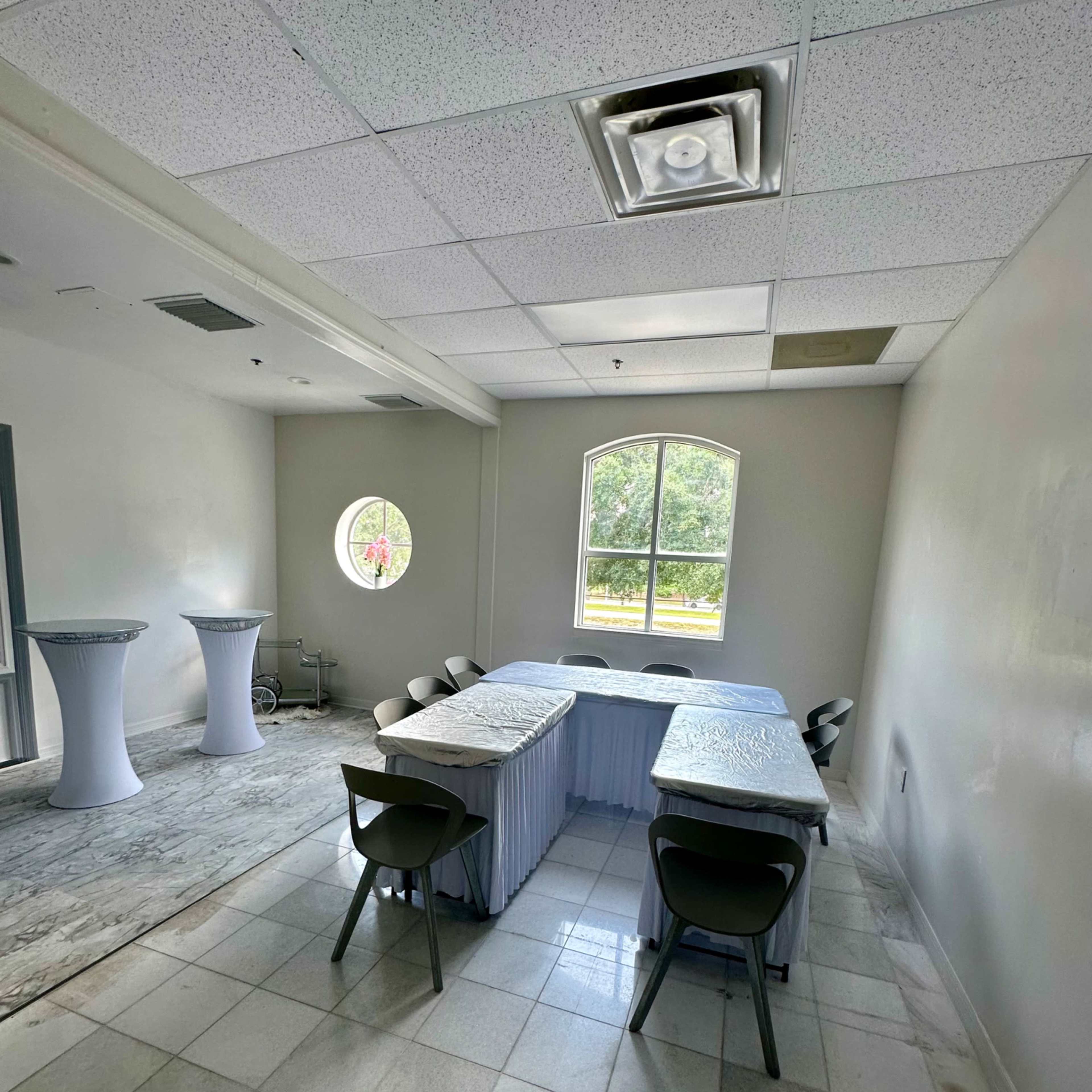 A small room with several tables arranged in a U-shape, surrounded by chairs, and featuring a circular window on one wall.