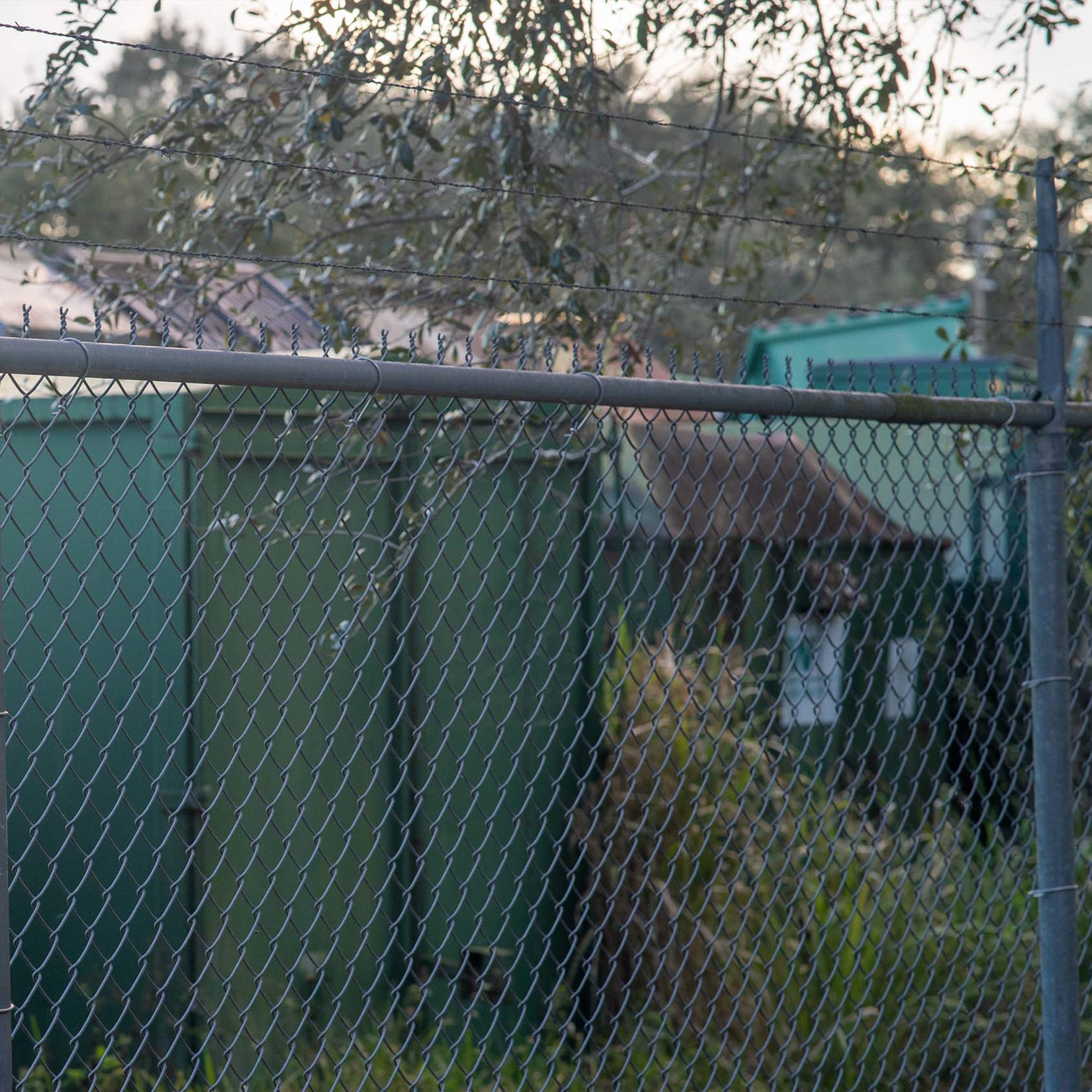 A chain-link fence enclosing several green storage containers amidst overgrown vegetation.